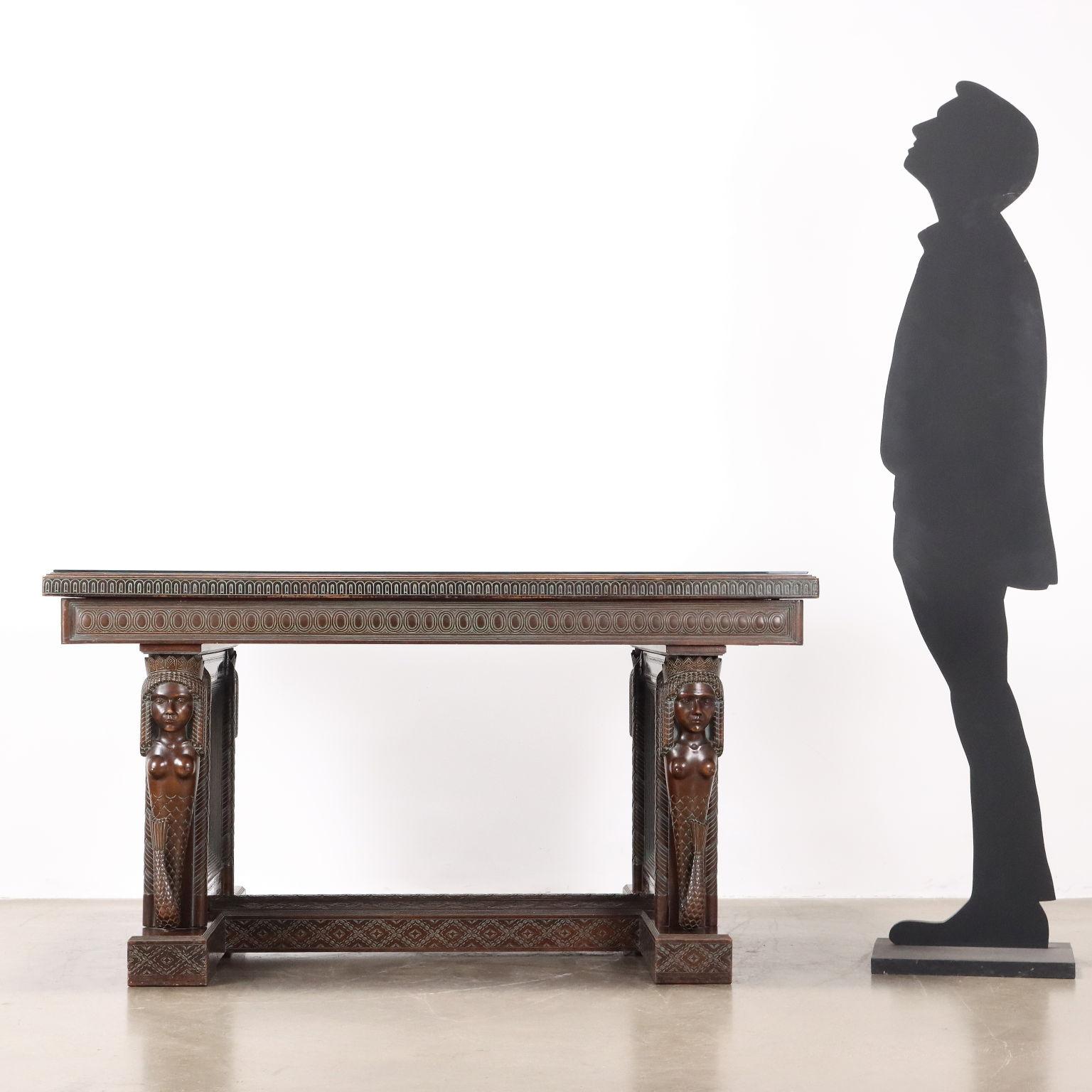 Mesa realizada por el artista Rino Galbiati, madera tallada en estilo étnico, dos pilares encerrados en dos sirenas talladas unidas por travesaños sostienen el tablero de la mesa, decorado con diversos motivos griegos en las bandas y en la parte