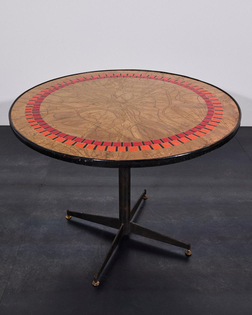 Une élégante table ronde caractérisée par un plateau en cuivre émaillé dont le centre présente des tons chauds avec des veines plus foncées, en contraste raffiné avec la décoration géométrique composée de deux cadres de carreaux rouge et orange vif.