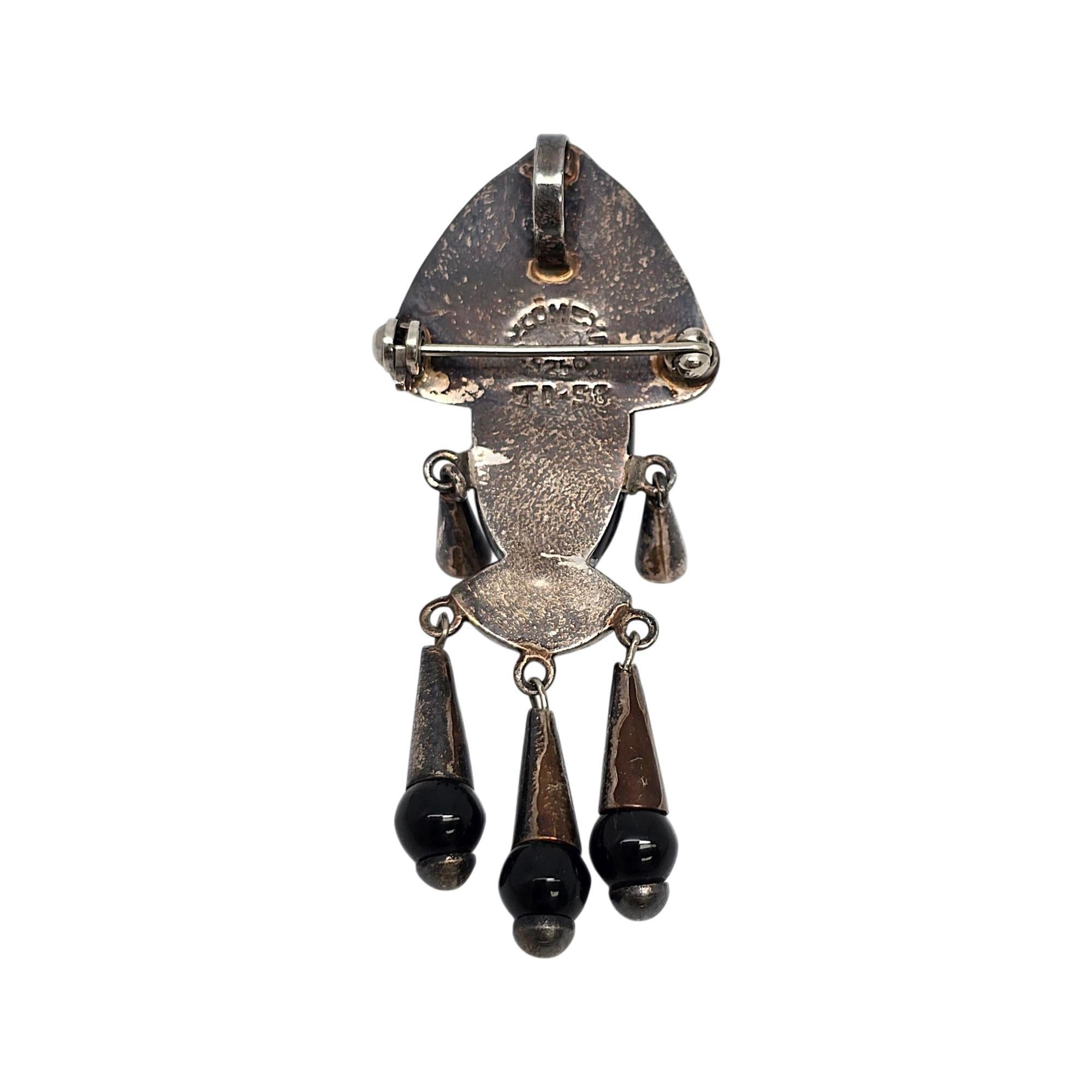 Spilla/pendente tribale di Taxco Mexico in argento sterling con volto di onice intagliato.

Questo pezzo presenta un volto in onice intagliato con coni pendenti, perline nere e pietre verdi incastonate. Può essere indossato come ciondolo o spilla.