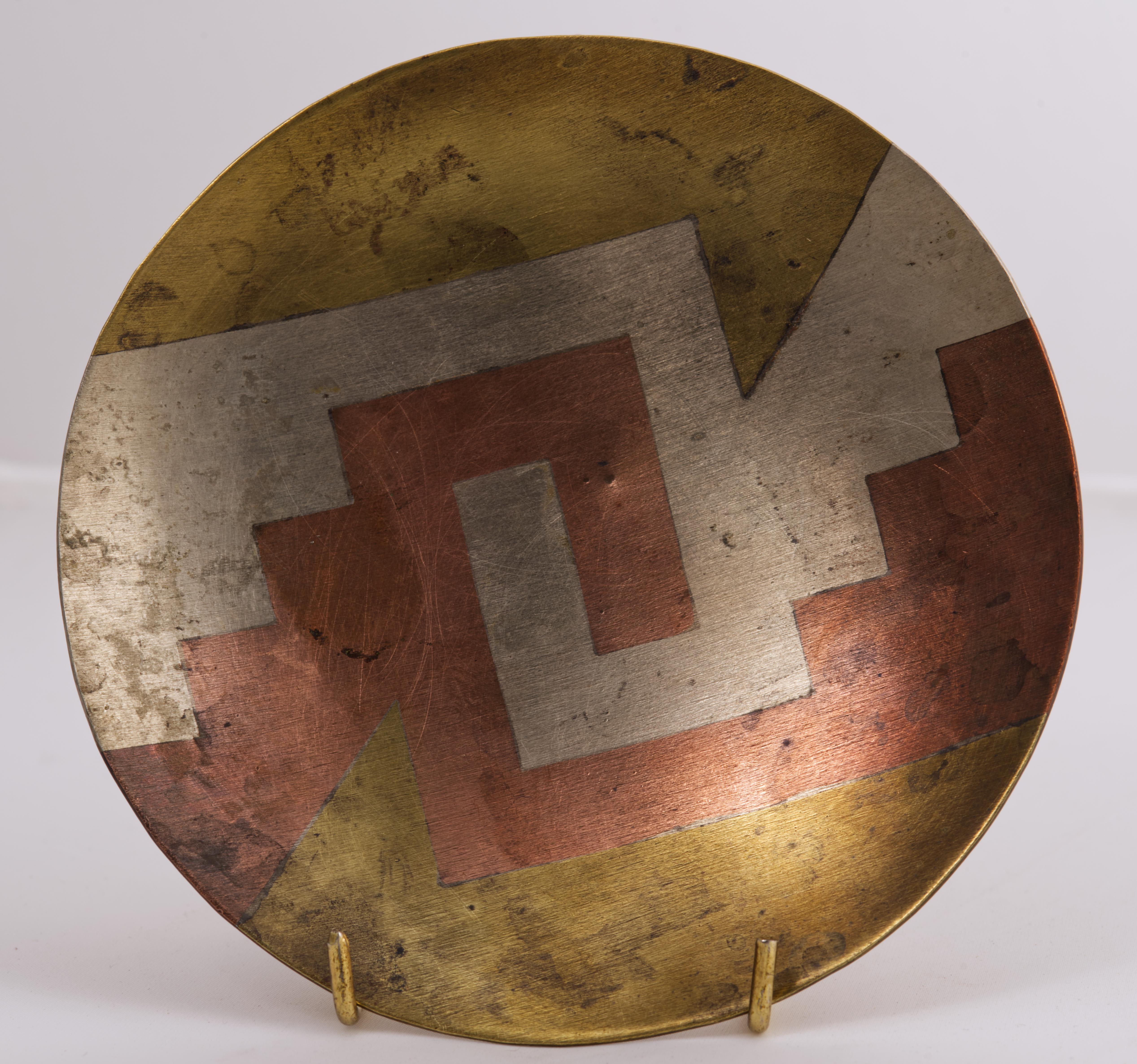 Il vassoio rotondo in metallo misto realizzato dagli argentieri messicani di Taxco, Messico, presenta un complesso motivo grafico modernista in ottone, rame, argento e bronzo. I metalli sono fusi senza soluzione di continuità in un'unica superficie