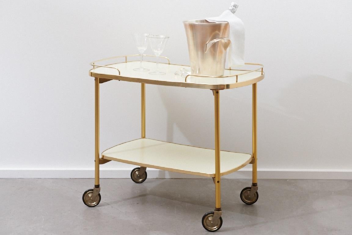 Restored: Serving trolley / bar cart Crema, 1950s , Formica surfaces in cream white, golden edging, polished brass rods, golden metal legs, on castors

Dimensions in cm:
Length 70
Width 42
Height 62.