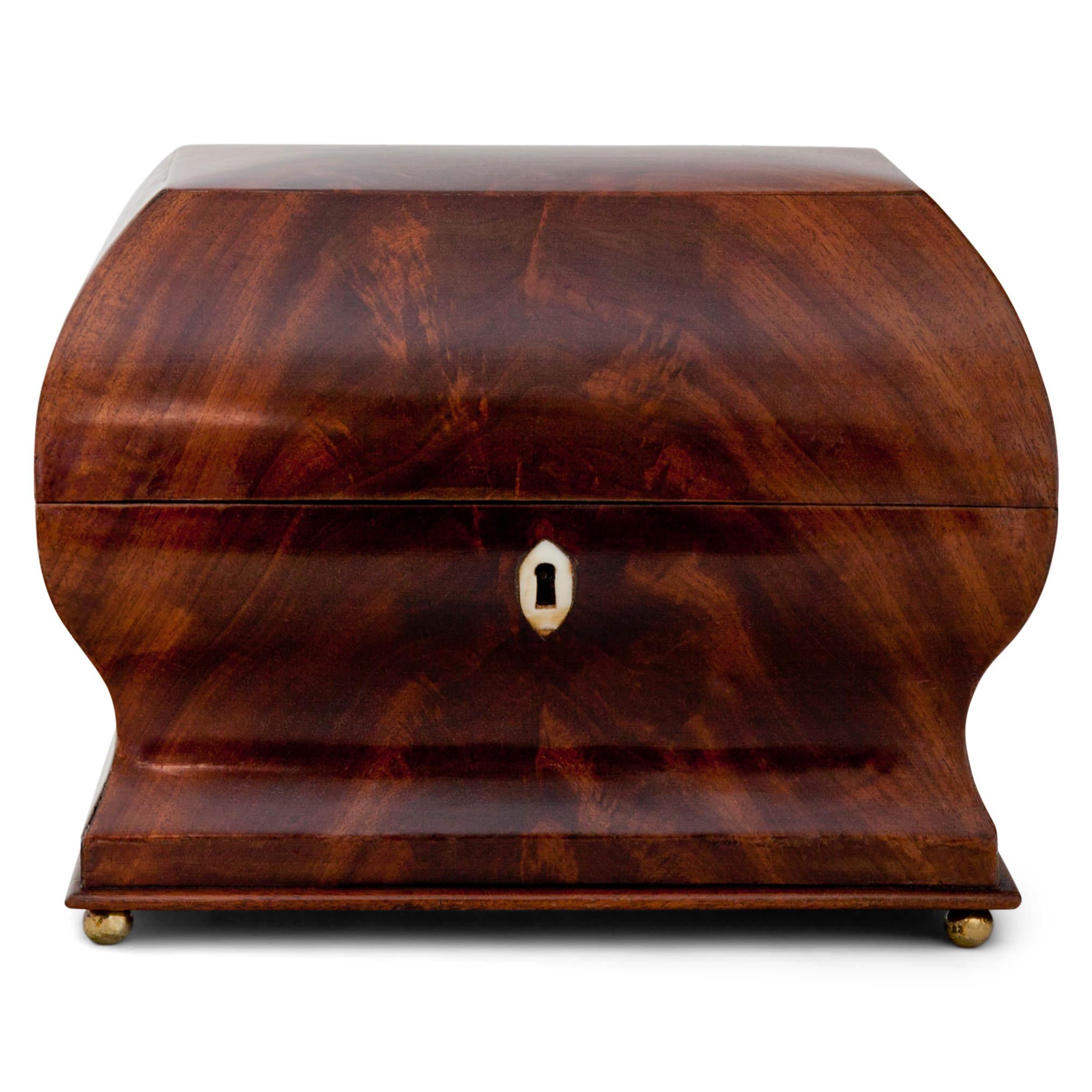 Lidded chest with a curved lid and a concave belly, standing on brass ball feet. The escutcheon is inlayed in bone. Mahogany veneered.