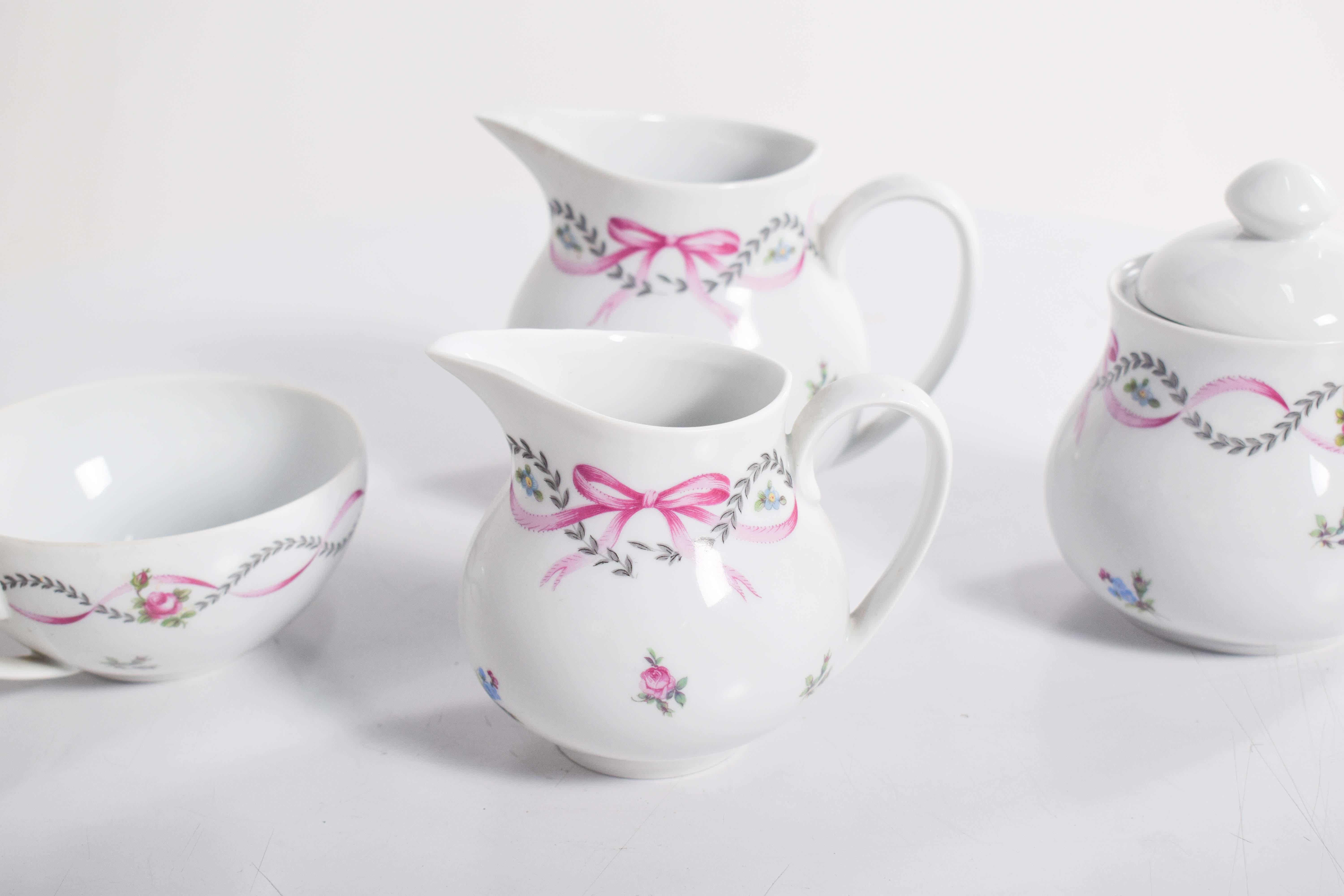 Questo affascinante servizio da tè, realizzato in pregiata porcellana di Limoges, presenta una delicata decorazione policroma con ghirlande floreali ed eleganti fiocchi rosa. Il design emana un'estetica romantica e senza tempo, con morbidi accenti