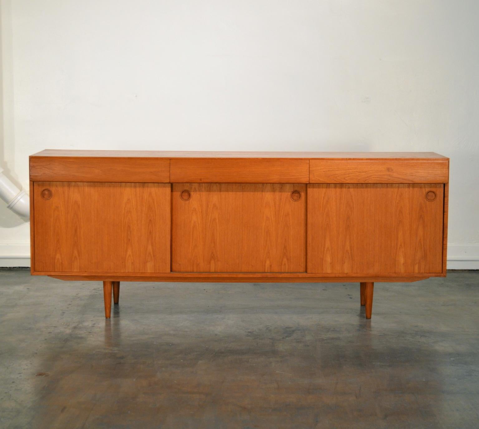 Stately 360 degree finished Teak Sideboard with three drawers on the top, having curved faces and hidden channel pulls. The three sliding front doors have expertly crafted inverted teak circular knobs. The top left drawer features a secondary