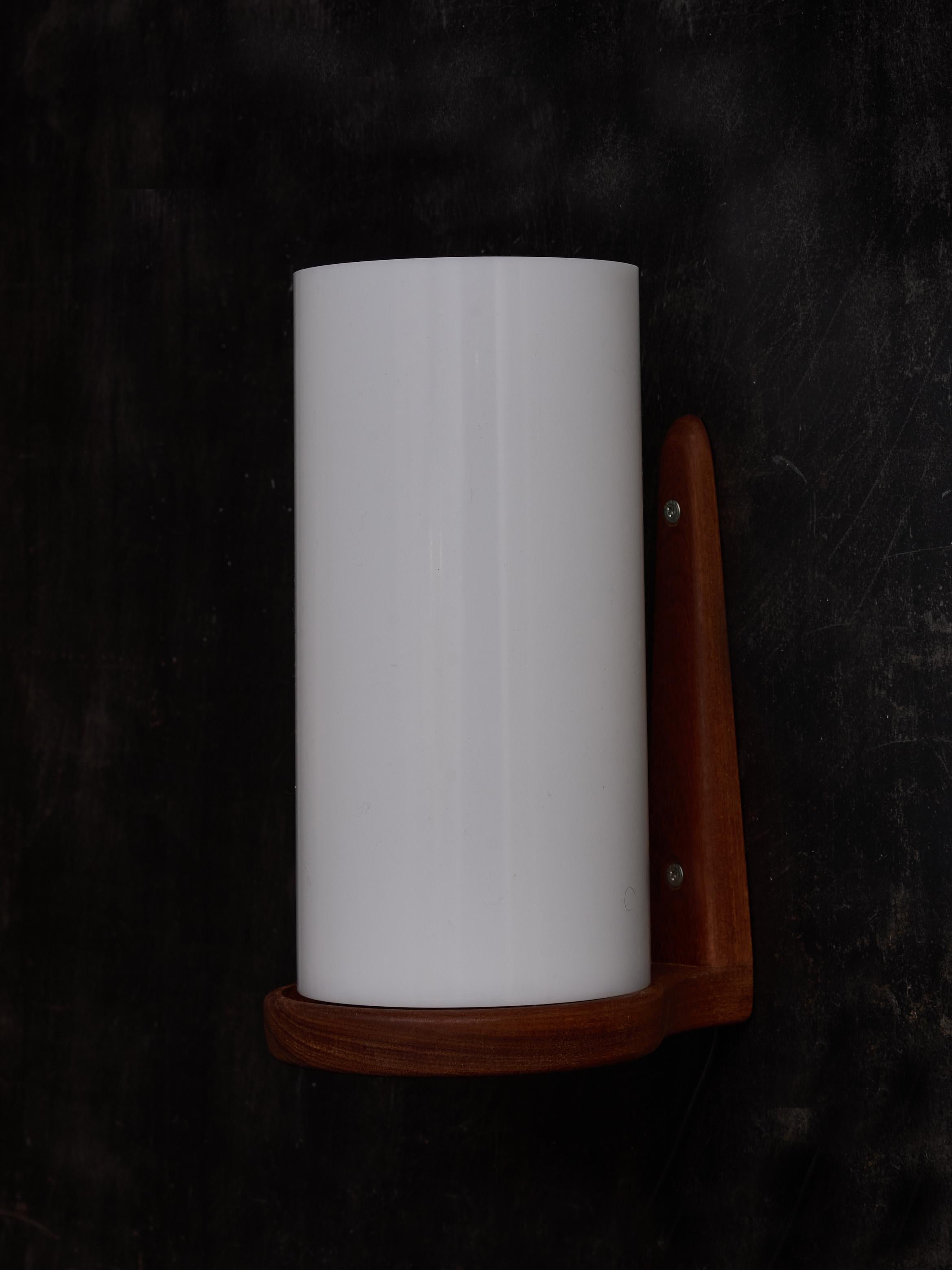 Applique da parete composte da una piastra posteriore in teak a forma di L che sostiene anche un paralume cilindrico in acrilico che diffonde la luce di una lampadina.

Grazie a un intaglio nella piastra posteriore, questi apparecchi possono essere