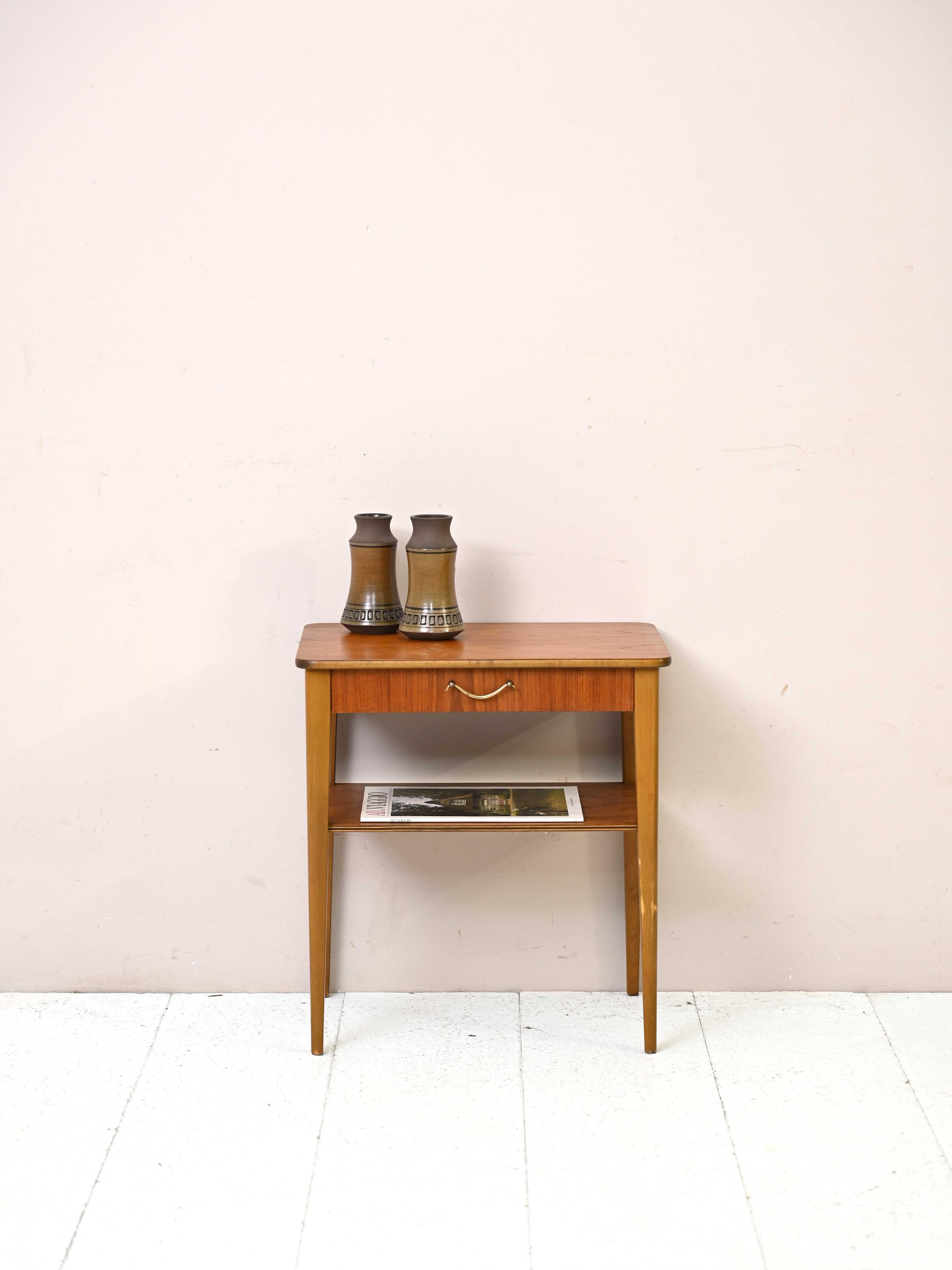 Scandinavian 1960s nightstand with metal handle.

A bedside table with the classic beauty of midcentury design.
The top with beveled corners is teak wood as is the drawer and magazine rack top. 
The tapered legs are made of birch.
The gilt
