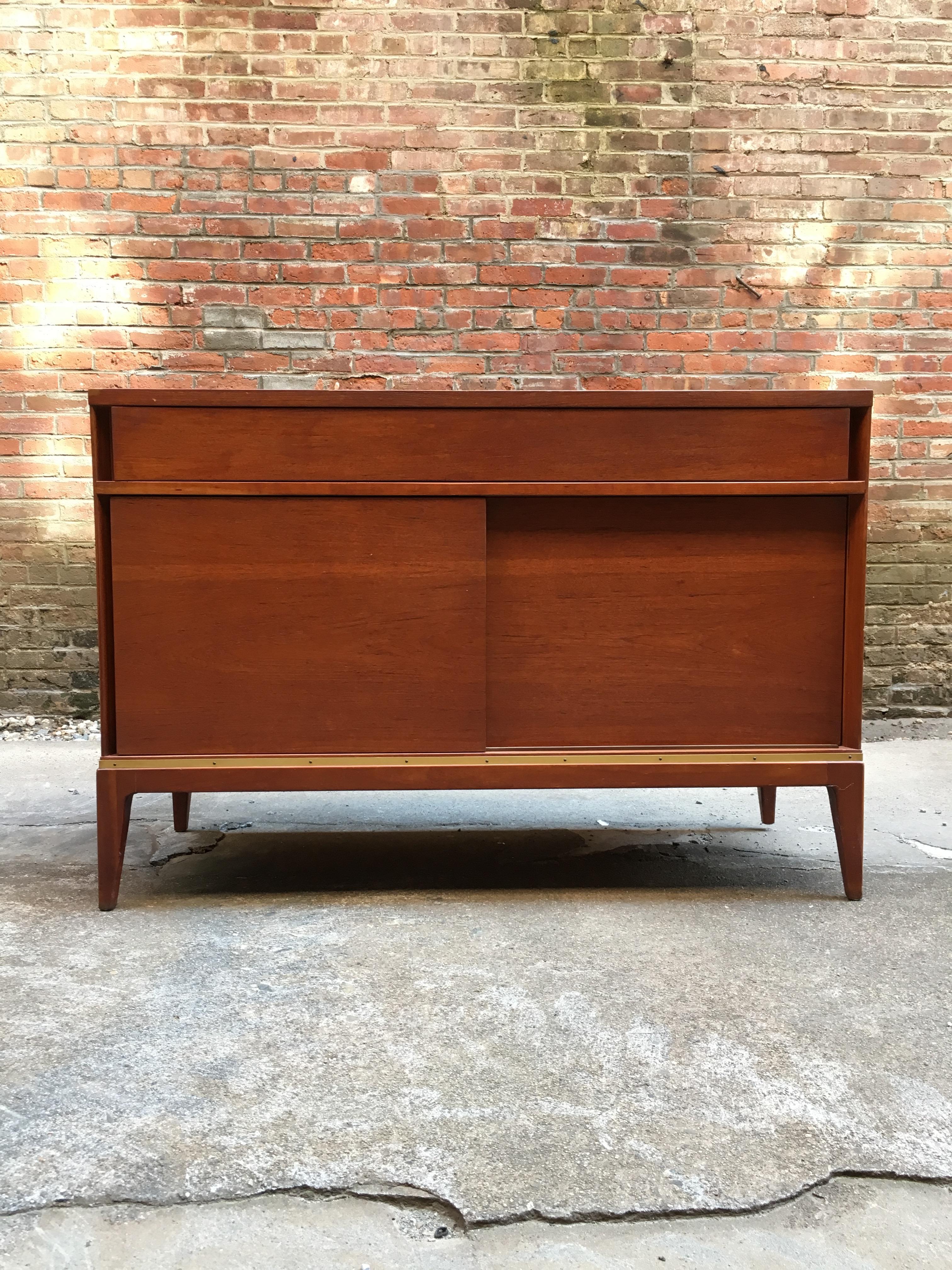 Teak with brass dressers retailed for John Stuart. True sign of quality when a piece of furniture has the John Stuart medallion in the top drawer. Warm teak with brass detail around the base. Featuring single top drawer over four hidden drawers,