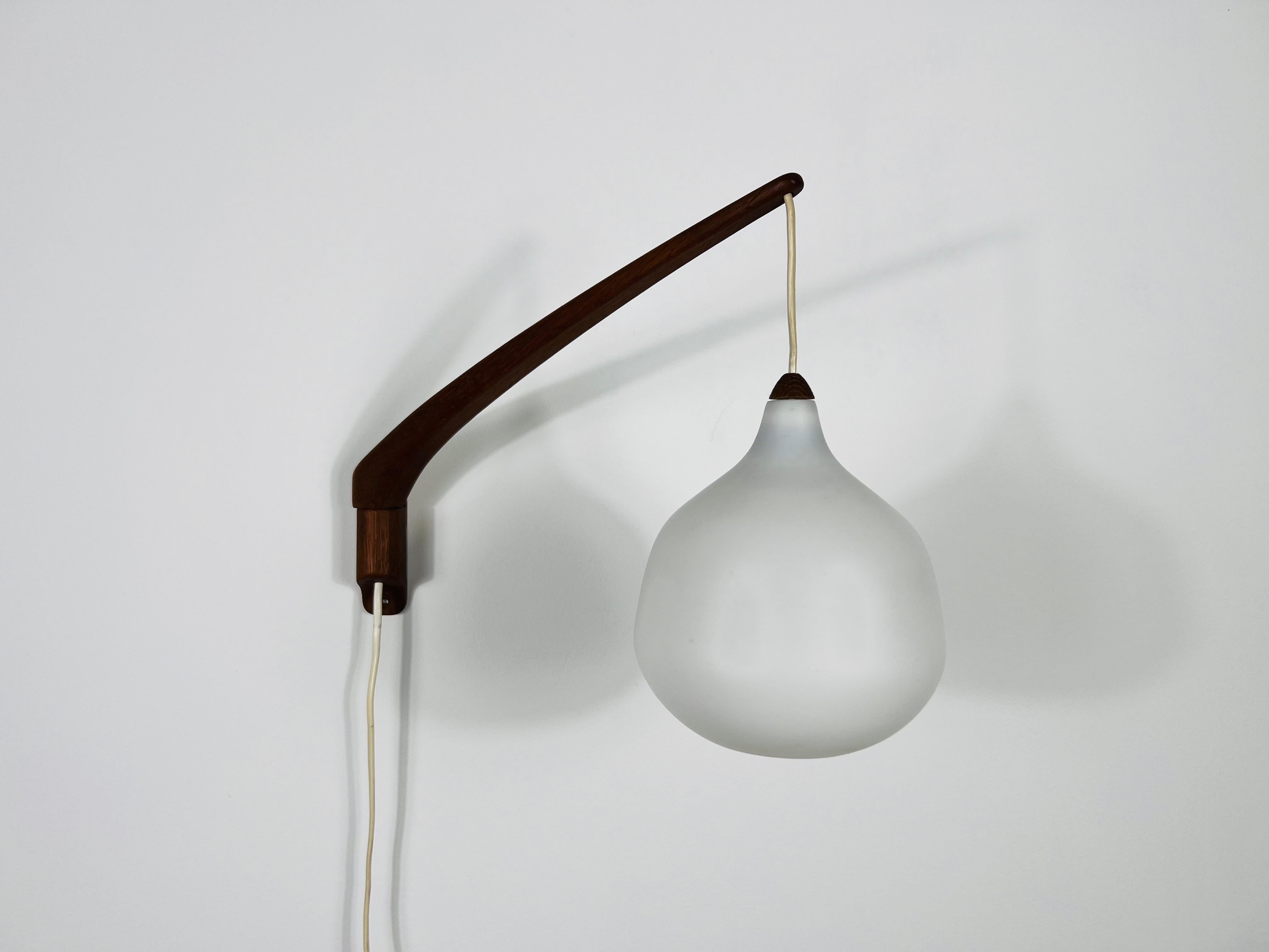 This elegant wall lamp, designed by Uno & Östen Kristiansson for Luxus Sweden in the 1960s, epitomizes mid-century modern design. It features a sleek teak arm and an opal glass shade, providing a sophisticated and high-quality aesthetic. The lamp is