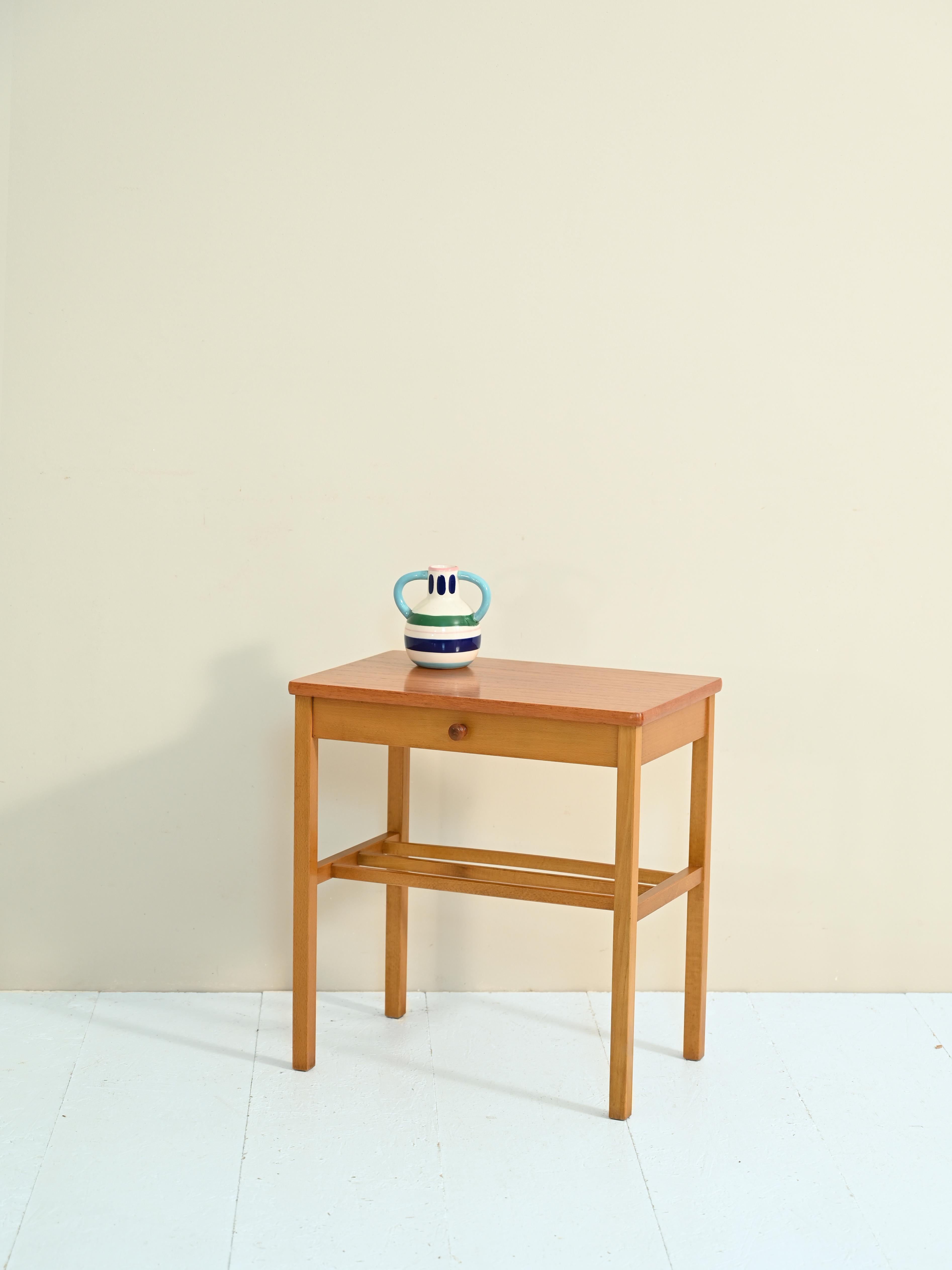 Teak bedside table with typical mid-century Scandinavian forms.

The stem is formed by a thin drawer supported by the long wooden legs.

This elegant bedside table is perfect for giving a northern European touch to a bedroom or can be placed at