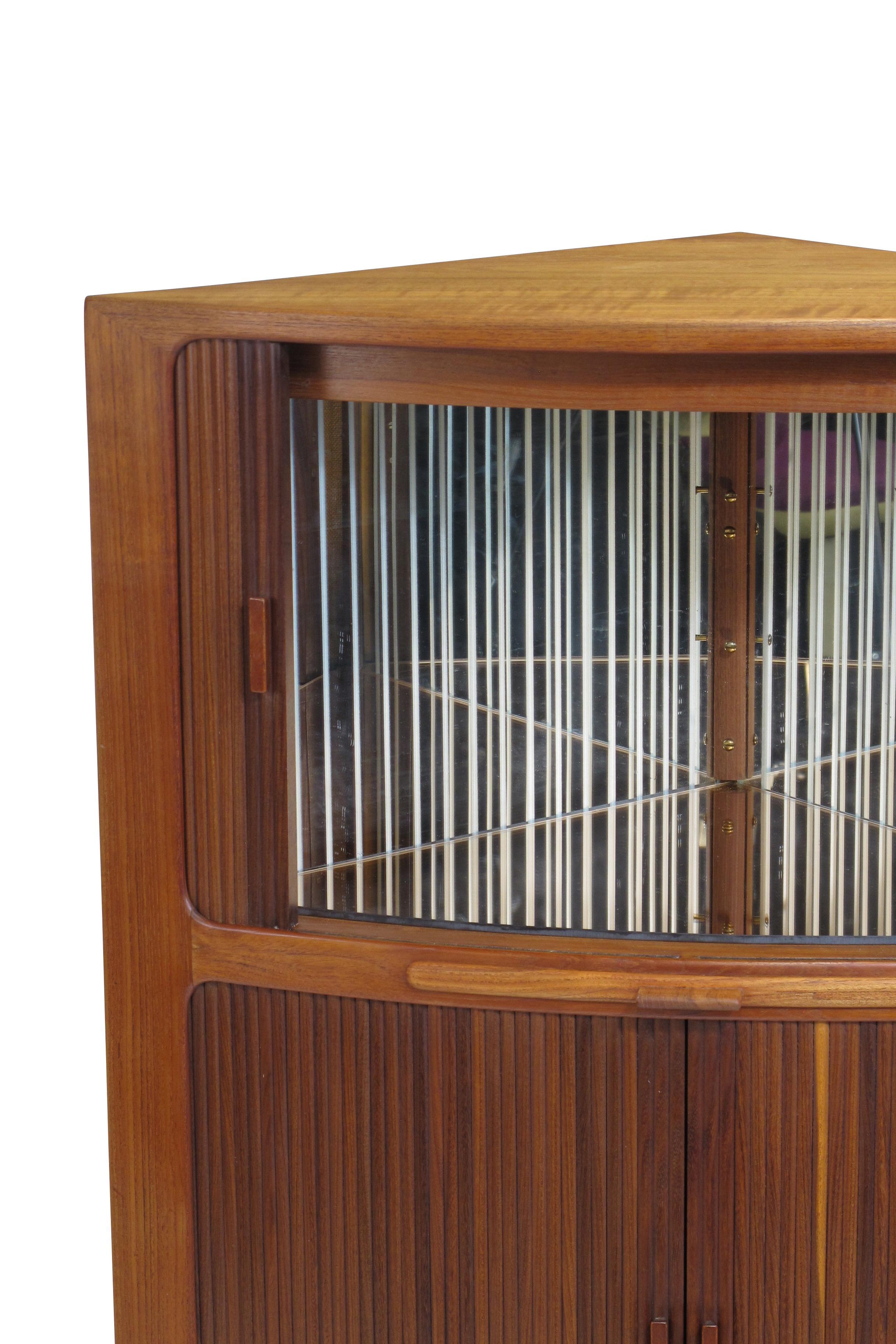 Teak corner cabinet attributed to Danish designer, Johannes Andersen. The cabinet is crafted of teak with two sections of tambour doors. The upper section reveals an interior with an etched mirror back, rose mirror base, and interior light. The