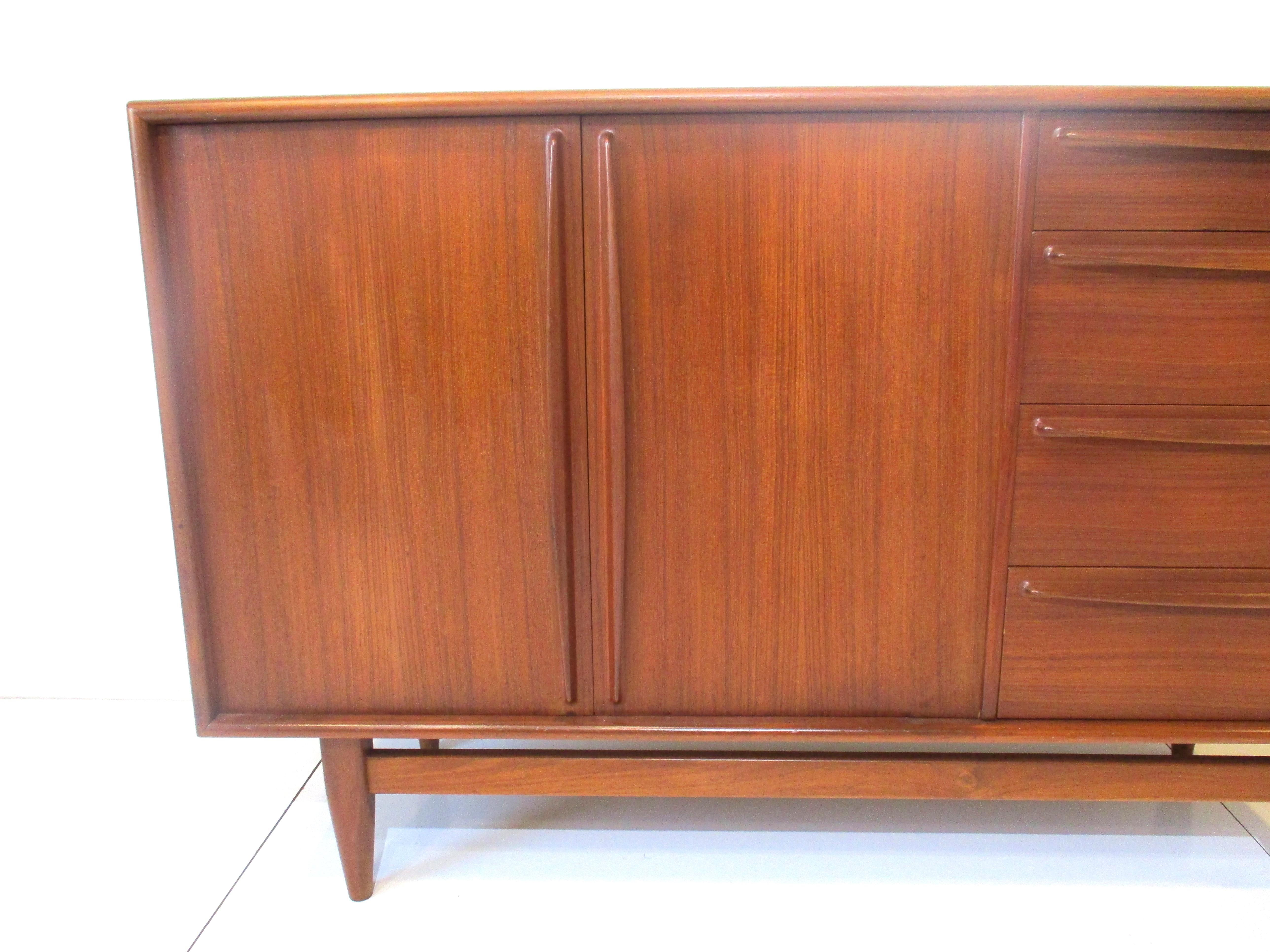 A very well crafted teak wood credenza with four drawers and two doors with long sleek matching handles and pulls . Having plenty of storage and two adjustable shelves inside the cabinet , all sitting on conical styled legs and cross stretchers