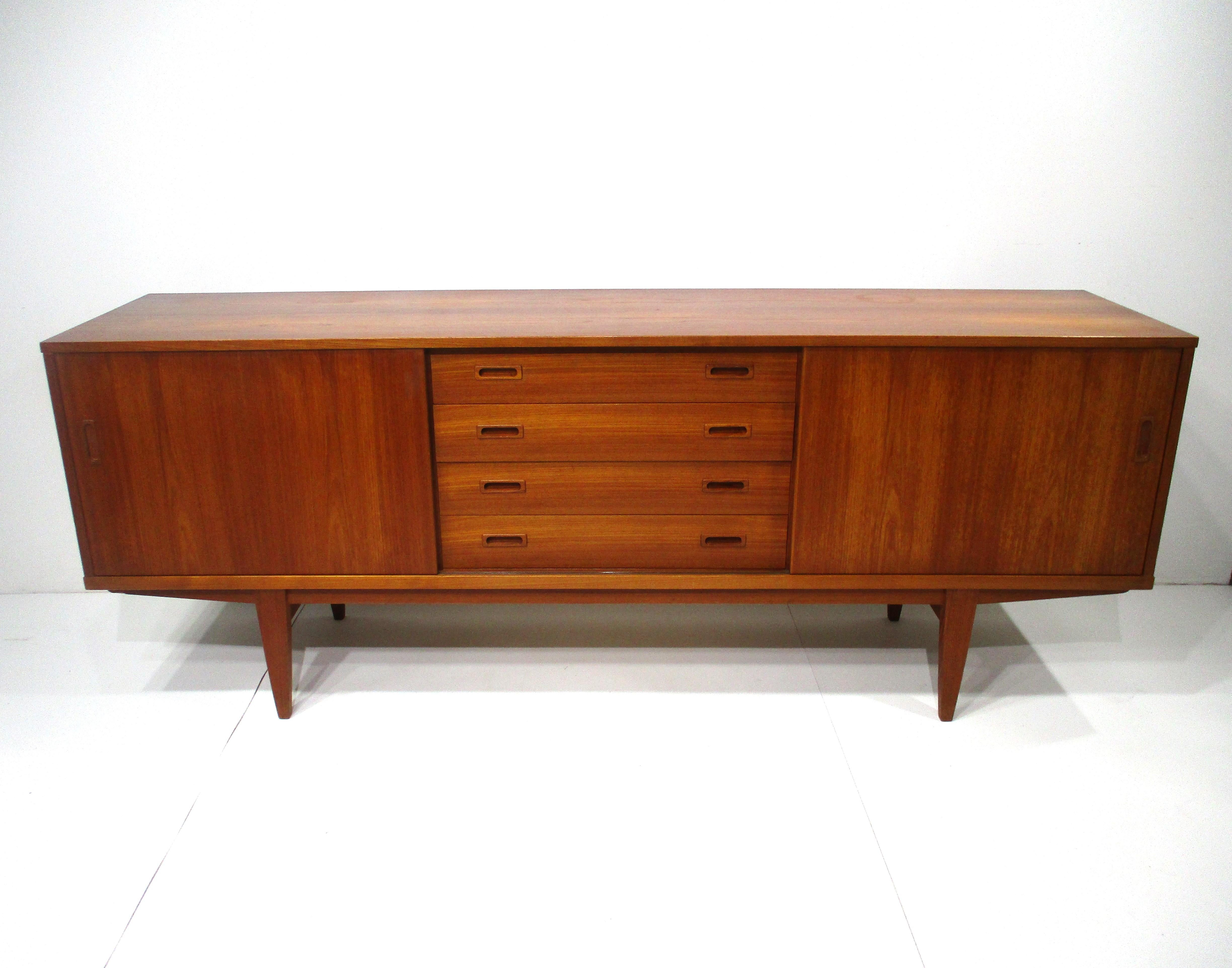 A very well crafted teak wood credenza sever with four drawers to the center the top one having a sliding tray for extra storage . The left side has two adjustable shelves and the other side has one non adjustable shelve with a cut out for taller