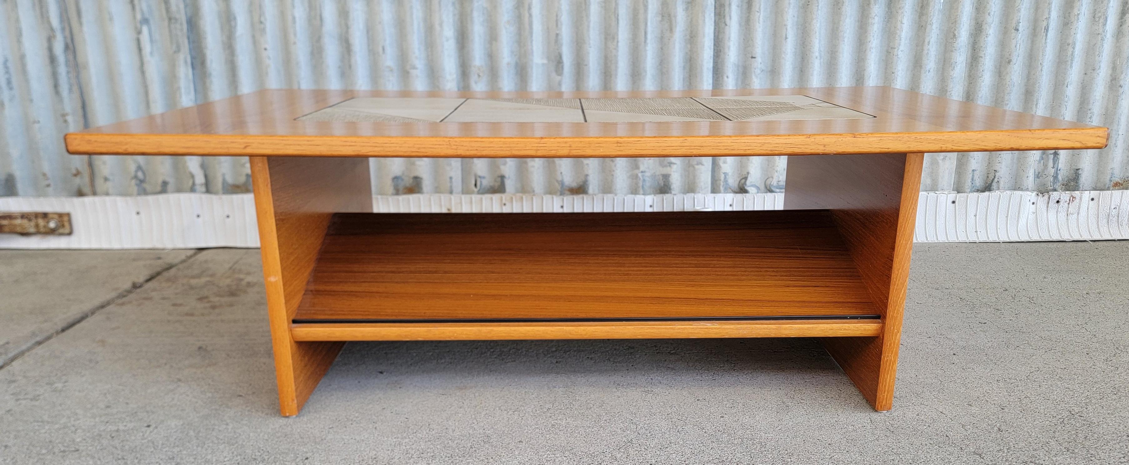 Teak Danish Modern coffee table with a geometric pattern ceramic tile top. Features an angled magazine or storage shelf underside. Circa. 1970's

48.5 inches wide.
29.5 inches deep.
16 inches high.