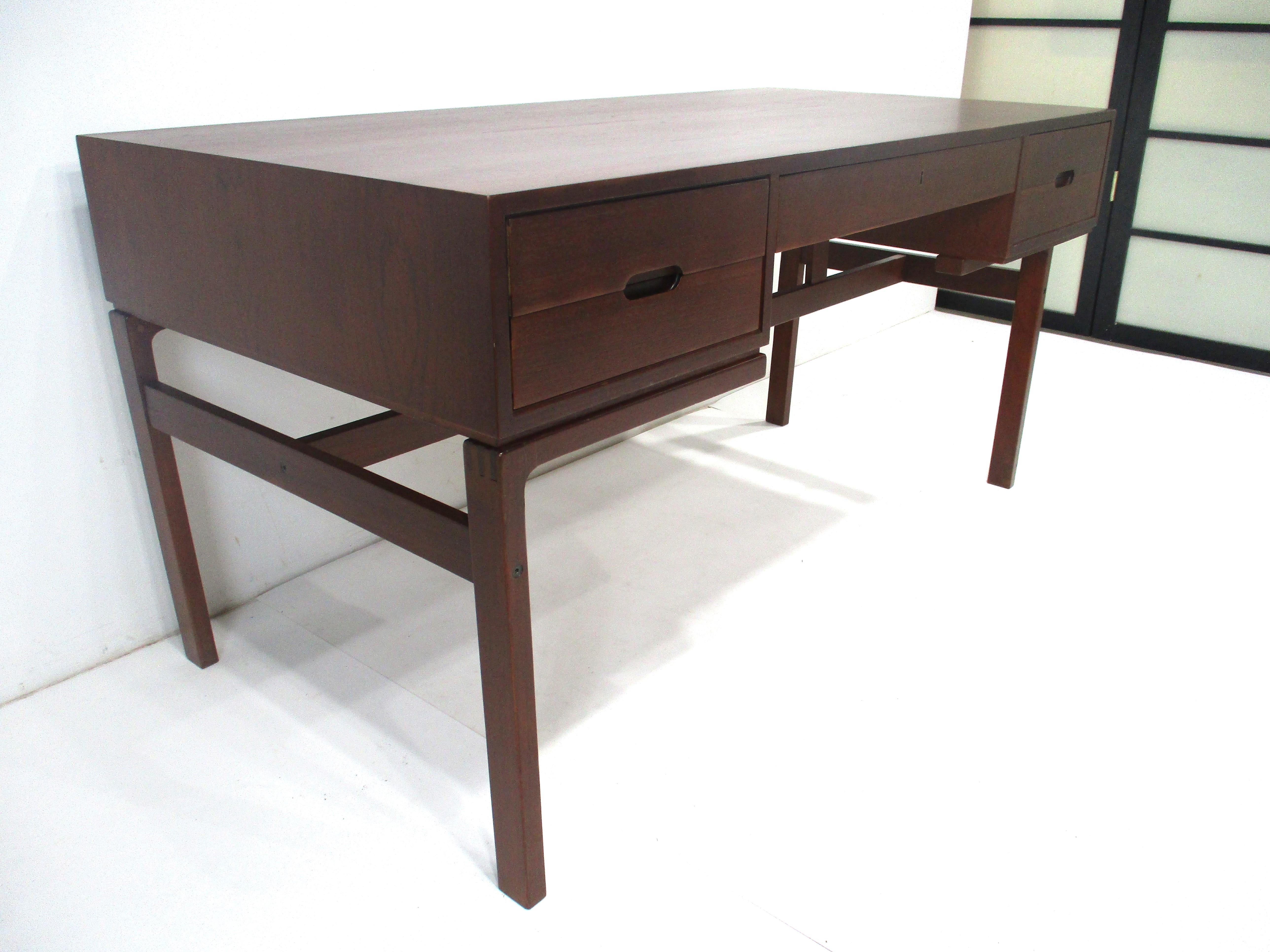 A dark teak desk almost in the tone and rich look of rosewood with two drawers to each side having inset pulls . The center has a slim drawer and the backside of the desk has two open storage areas and H type legs but one side has a shorter width or