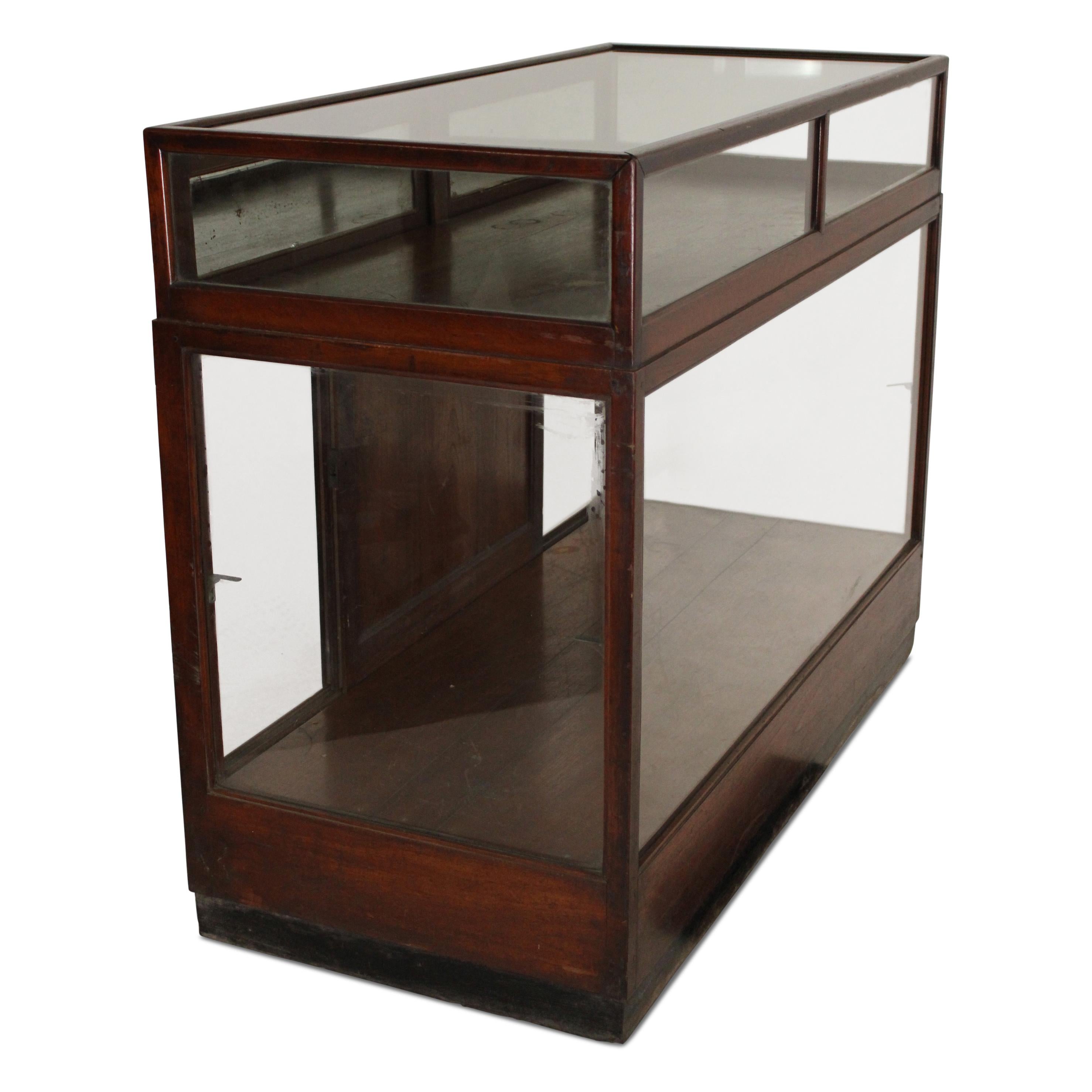 A teak shop display counter cabinet featuring a glass top and glass panels on three sides, along with a pair of sliding door panels below. The counter also includes two drawers at the bottom for added storage. It retains its original polish,