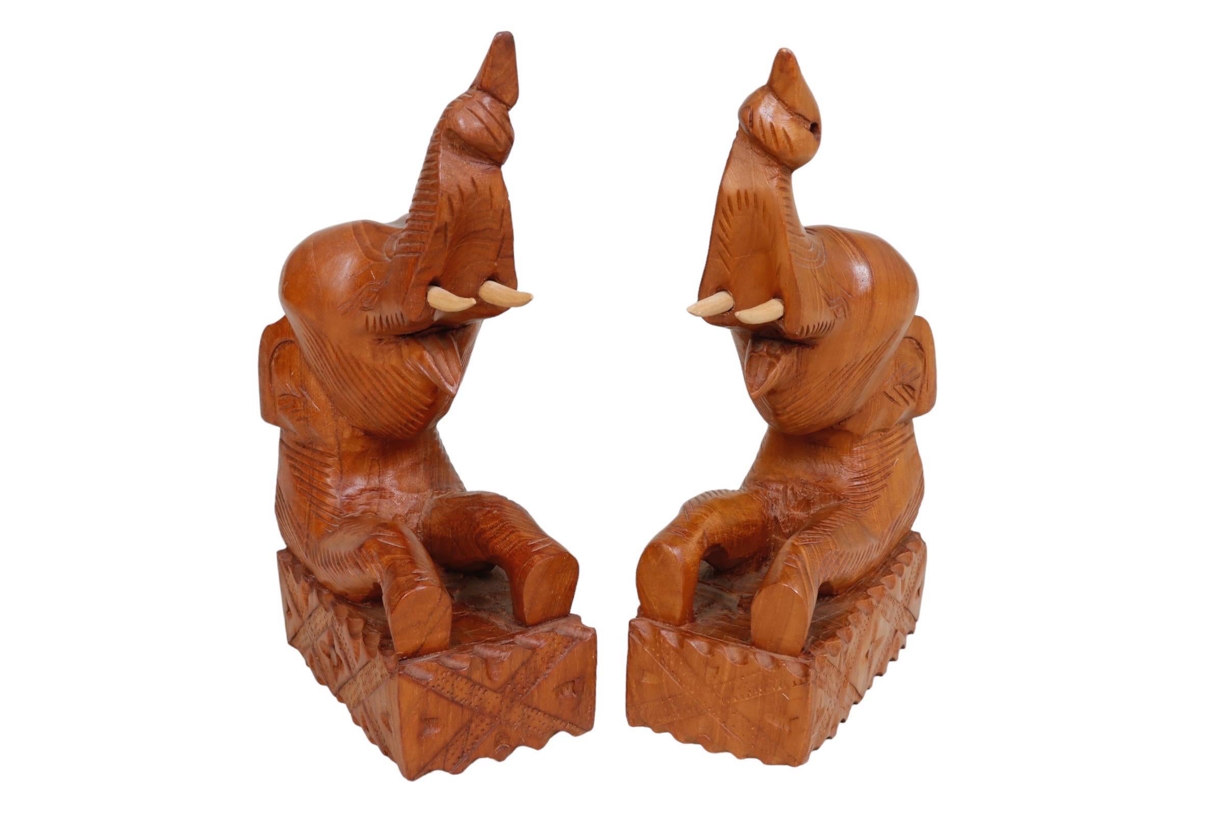 A pair of Asian elephant bookends made of teak. Hand carved with trunks raised triumphantly, used to signify good fortune, positive energy, and prosperity. Simple lines define the features of the elephant including eyes, ears and skin. Front legs