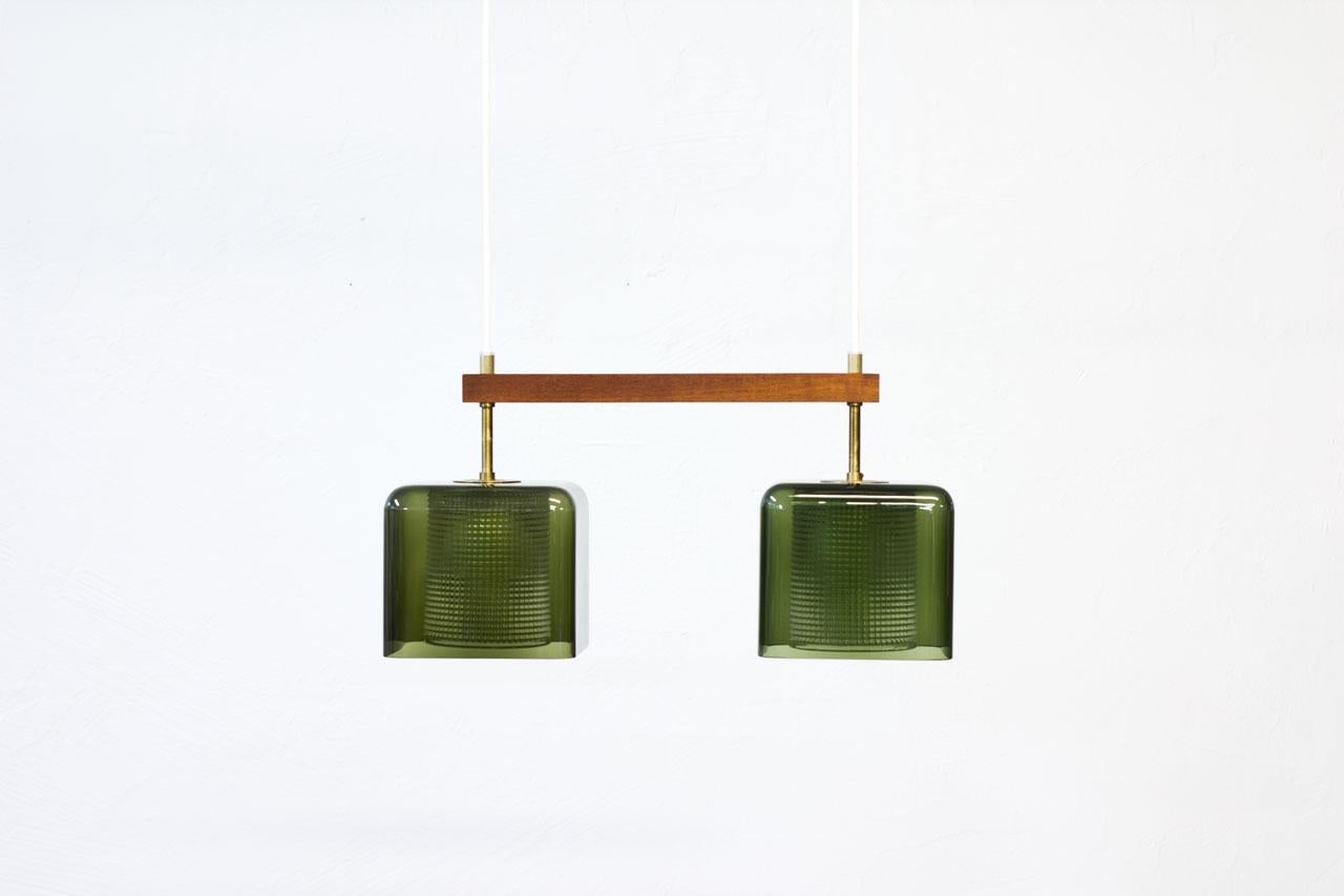 Pendant lamp designed by Carl Fagerlund for Swedish glass company Orrefors during the 1960s. Square shaped cups in green tinted glass with internal clear pressed glass diffuser, framed by a teak stretcher. Brass fittings with original brass ceiling