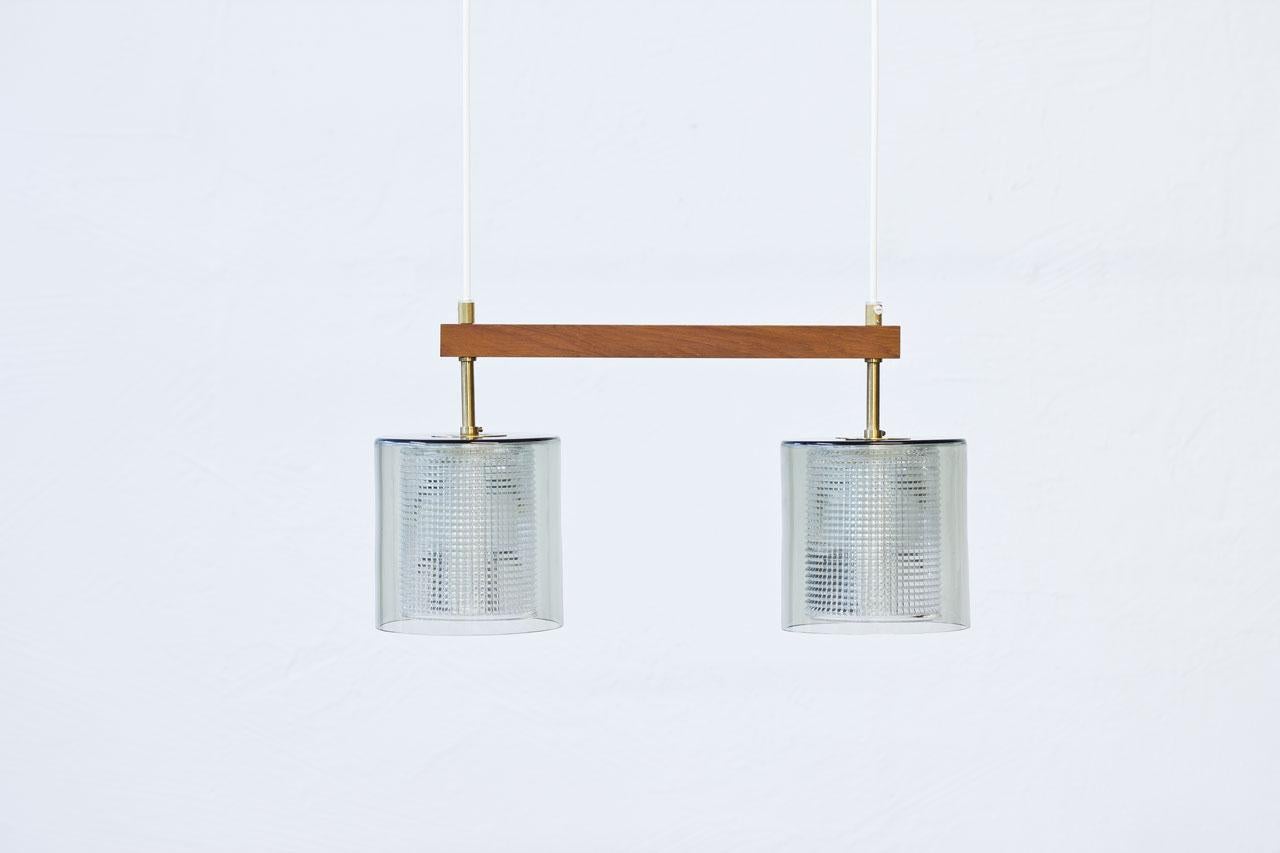 Pendant lamp designed by Carl Fagerlund for Swedish glass company Orrefors during the 1960s. Cylindrical cups in clear grey tinted glass with internal clear pressed glass diffuser, framed by a teak stretcher. Brass fittings with original brass