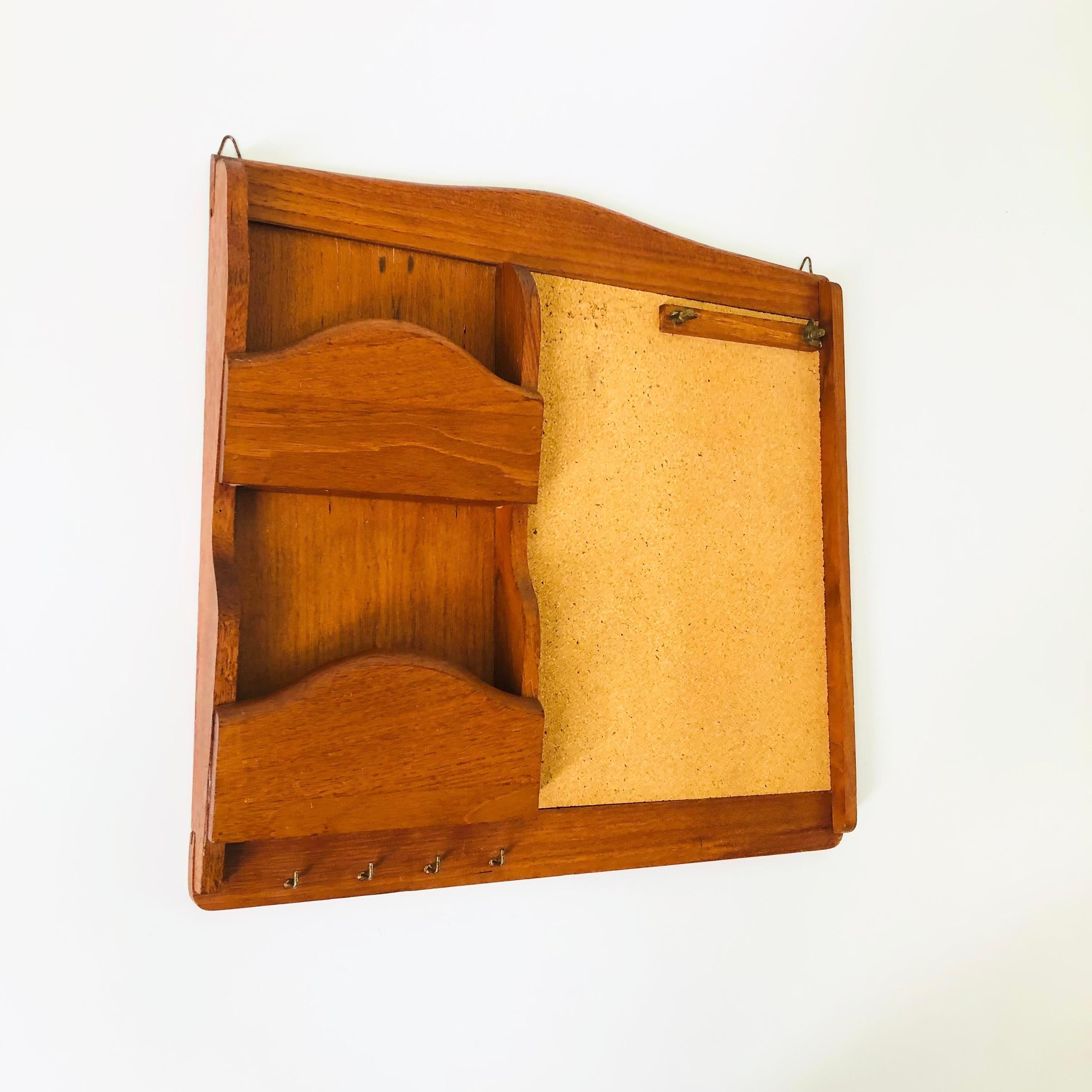 A vintage teak letter holder and cork board. There are 2 compartments for storing paper items with hooks below for hanging keys. A rectangular cork board is on the right with screws for clamping a note pad. A great entry way organizational piece.

