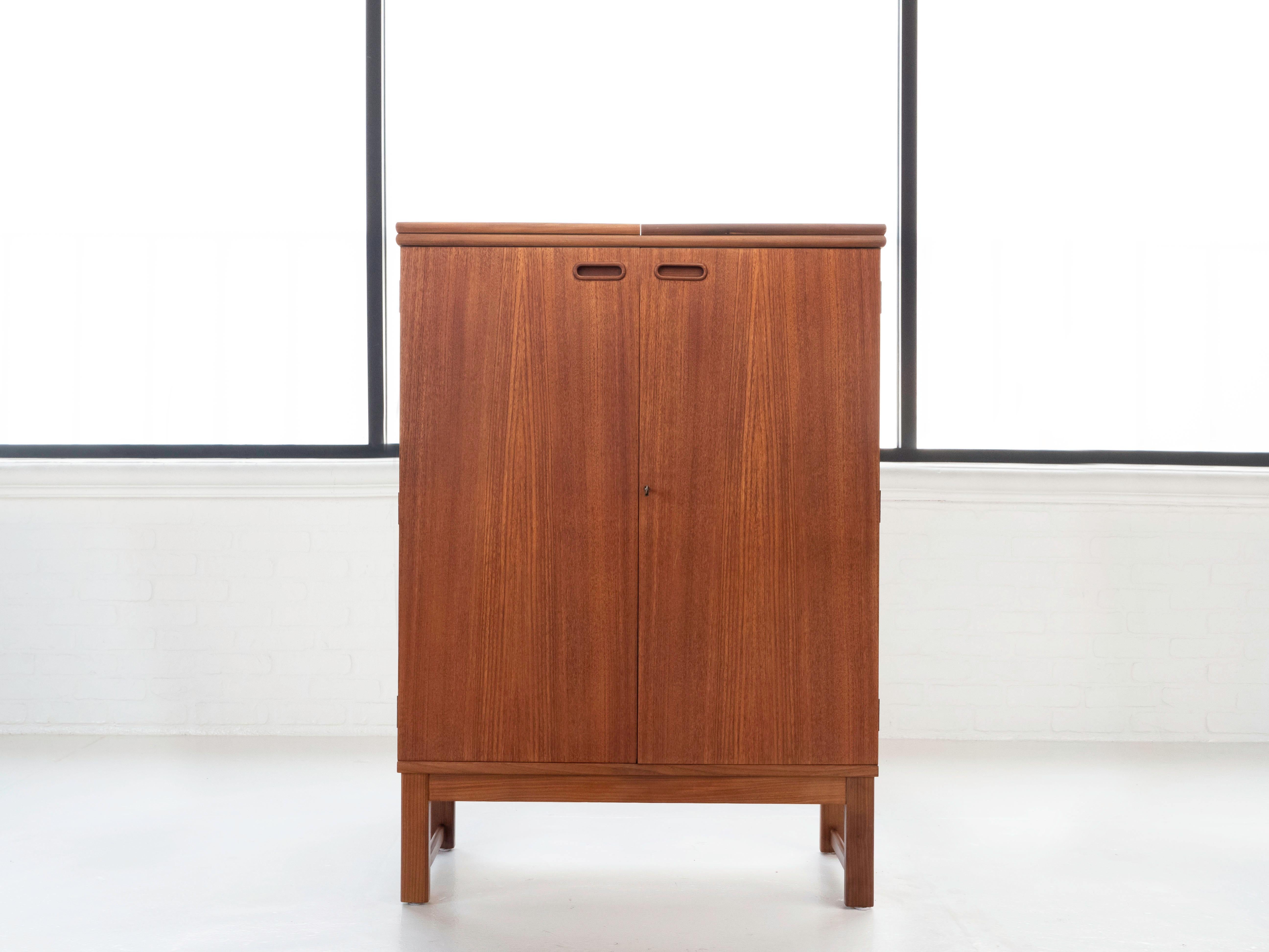 Teak mid-century dry bar cabinet by maker Turnbridge of London.  Circa 1960's.  The cabinet has a two-door front that opens to a bar shelf with a mirrored back.  There is a flip top.  When the top is flipped, there is a faux teak laminate that you