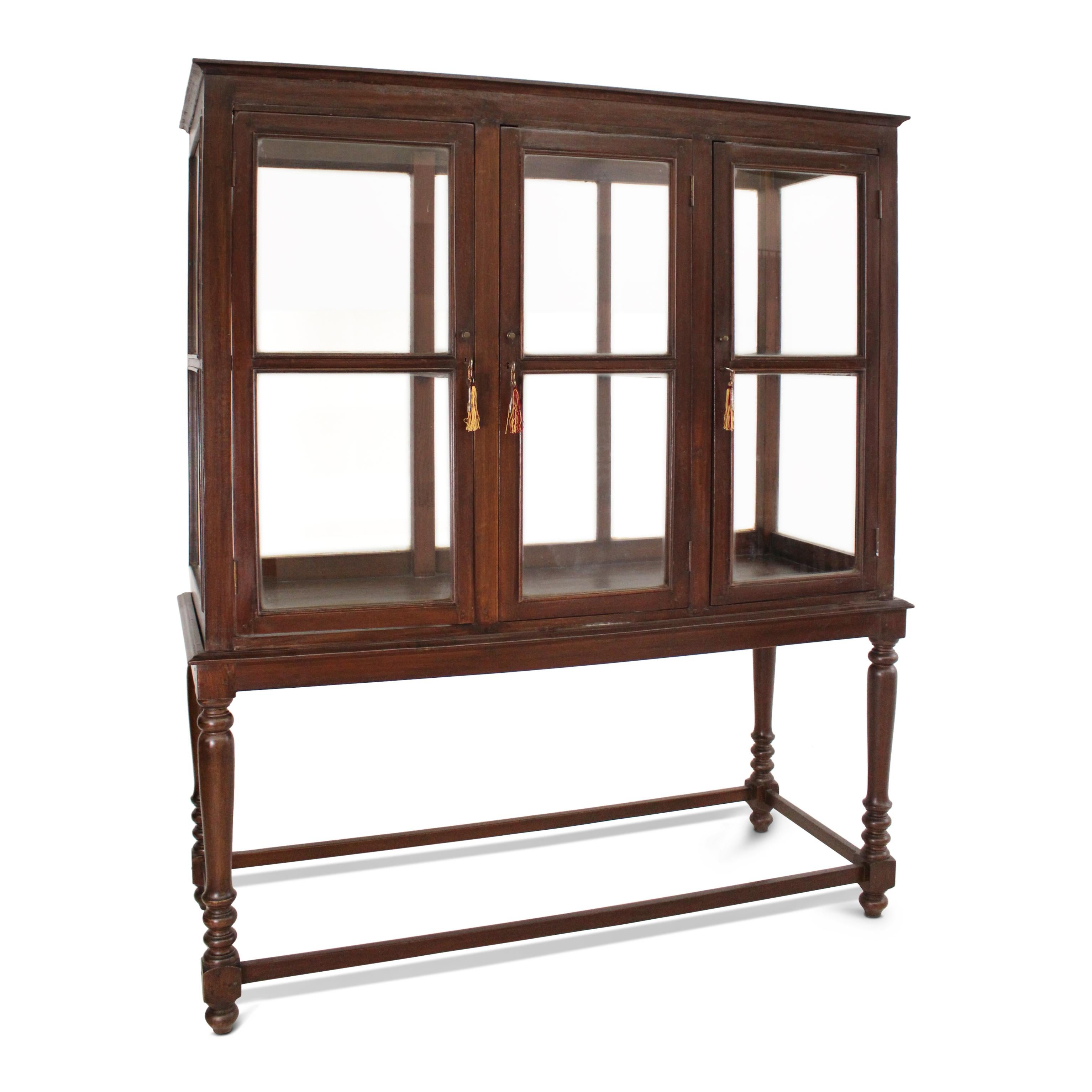 A teak museum vitrine cabinet, in excellent condition. This generously sized cabinet features glass panels on all sides, three front doors, and glass shelving inside.