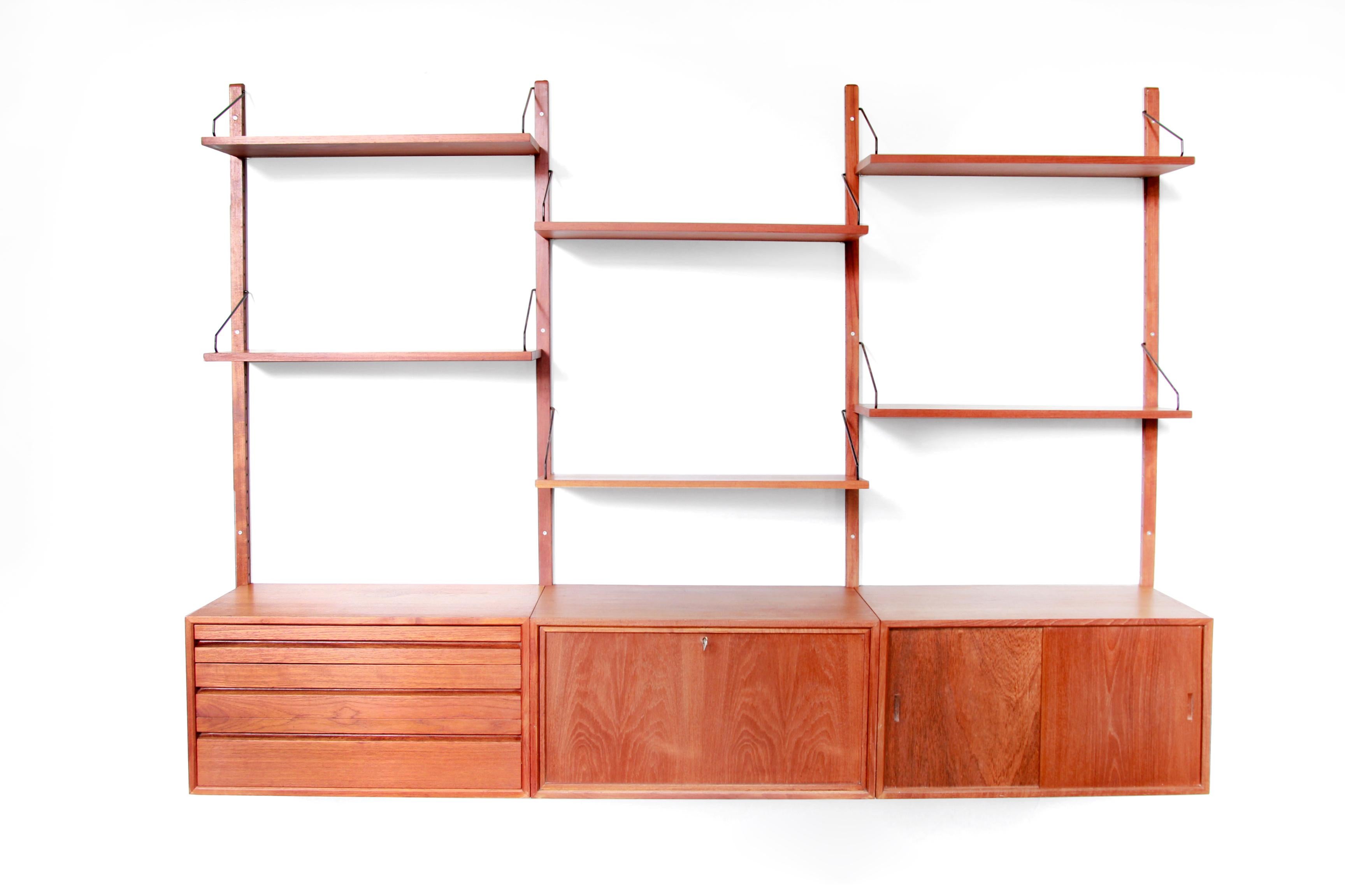 Teak wooden wall system designed by Poul Cadovius and produced by Royal System Denmark. This wall cabinet from the sixties consists of 4 wooden uprights, 6 shelves, a cabinet with two sliding doors, a cabinet with a flap that can be used as a bar