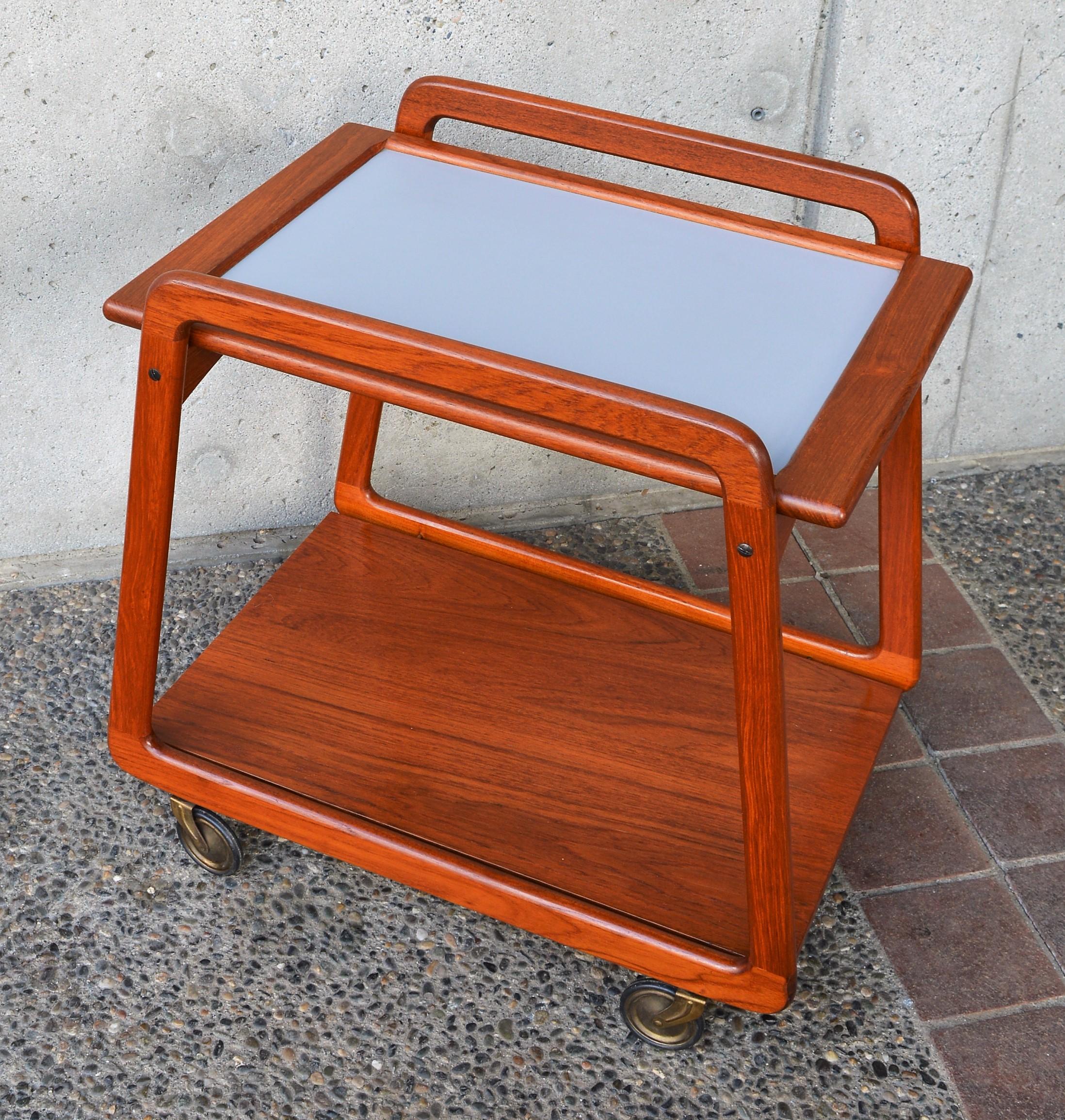 This super fun Danish modern teak bar cart or tea trolley has awesome lines! It features trapezoidal shaped sides, arced upper braces that support the two side’s tray top, a lower floating shelf held by brass pins, and Classic brass skinny 1950s