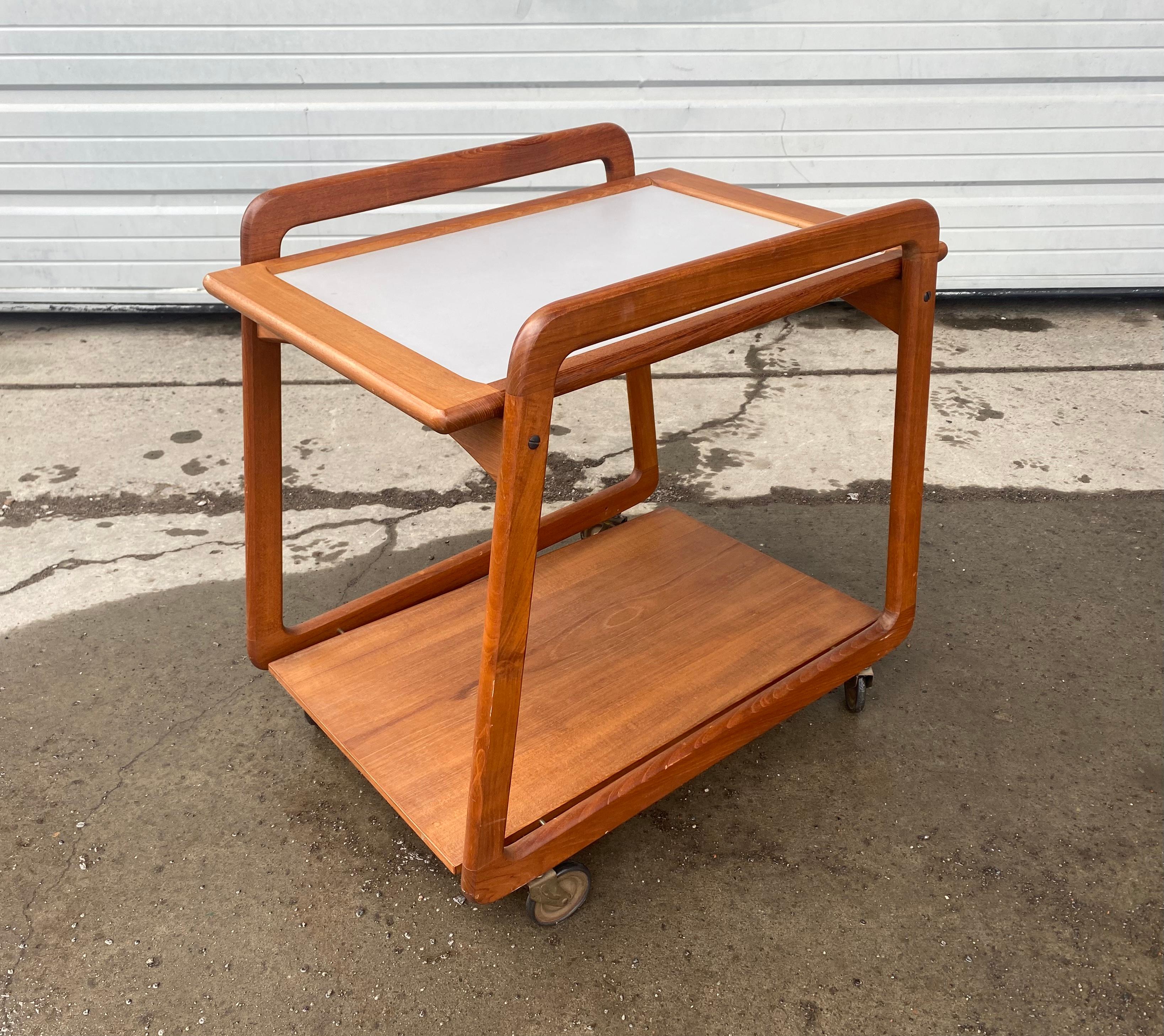 Danish modern teak bar cart or tea trolley has awesome lines! It features trapezoidal shaped sides, arced upper braces that support the two side’s tray top, a lower floating shelf held by brass pins, and Classic brass skinny 50s casters. The
