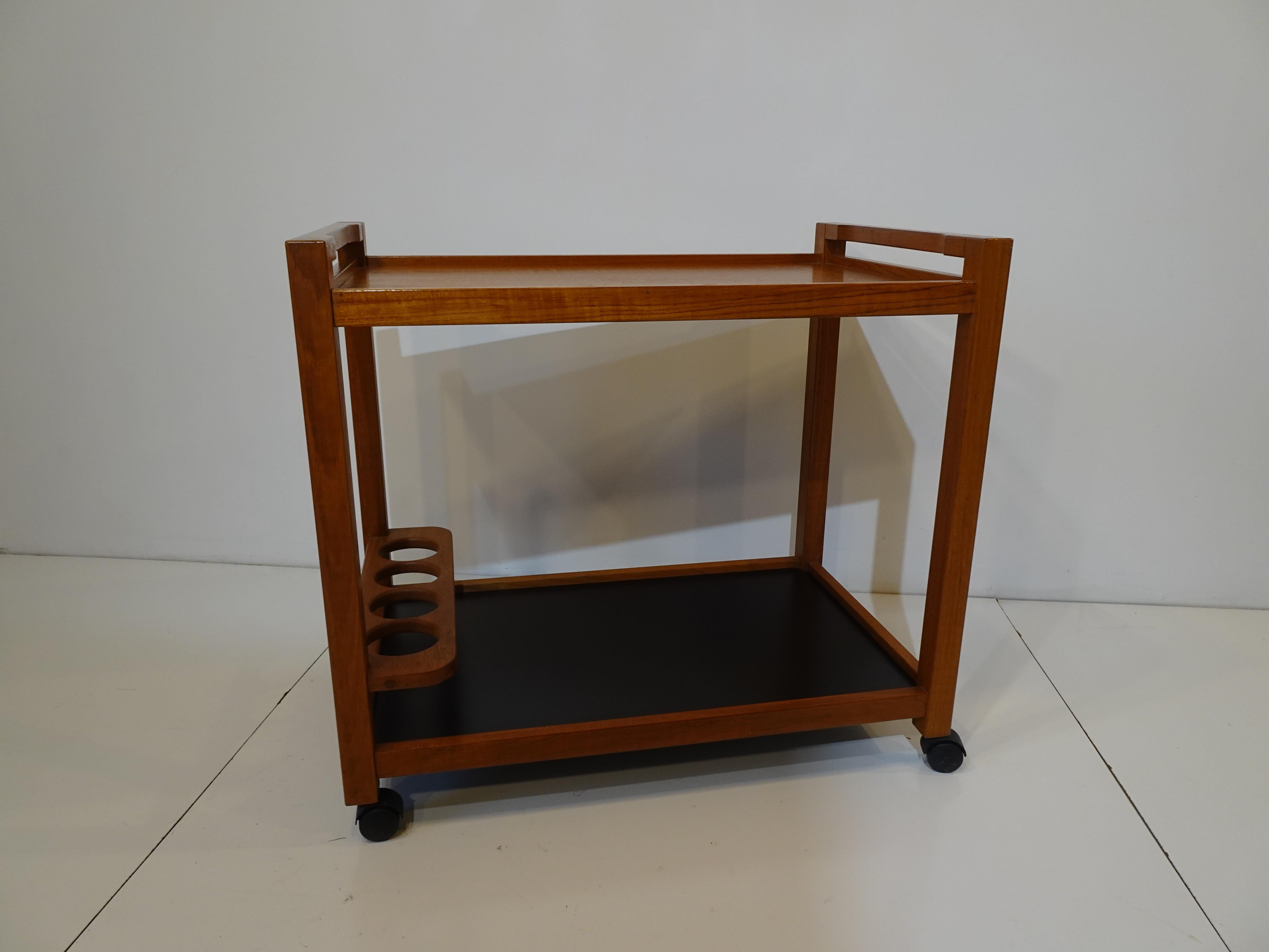 A two tiered rolling bar trolley / cart with the top in teak having handles to each side, the bottom is in a satin black laminate with a teak four bottle holder to one end. Designed in the manner of Eric Buch and Danish Modern.
