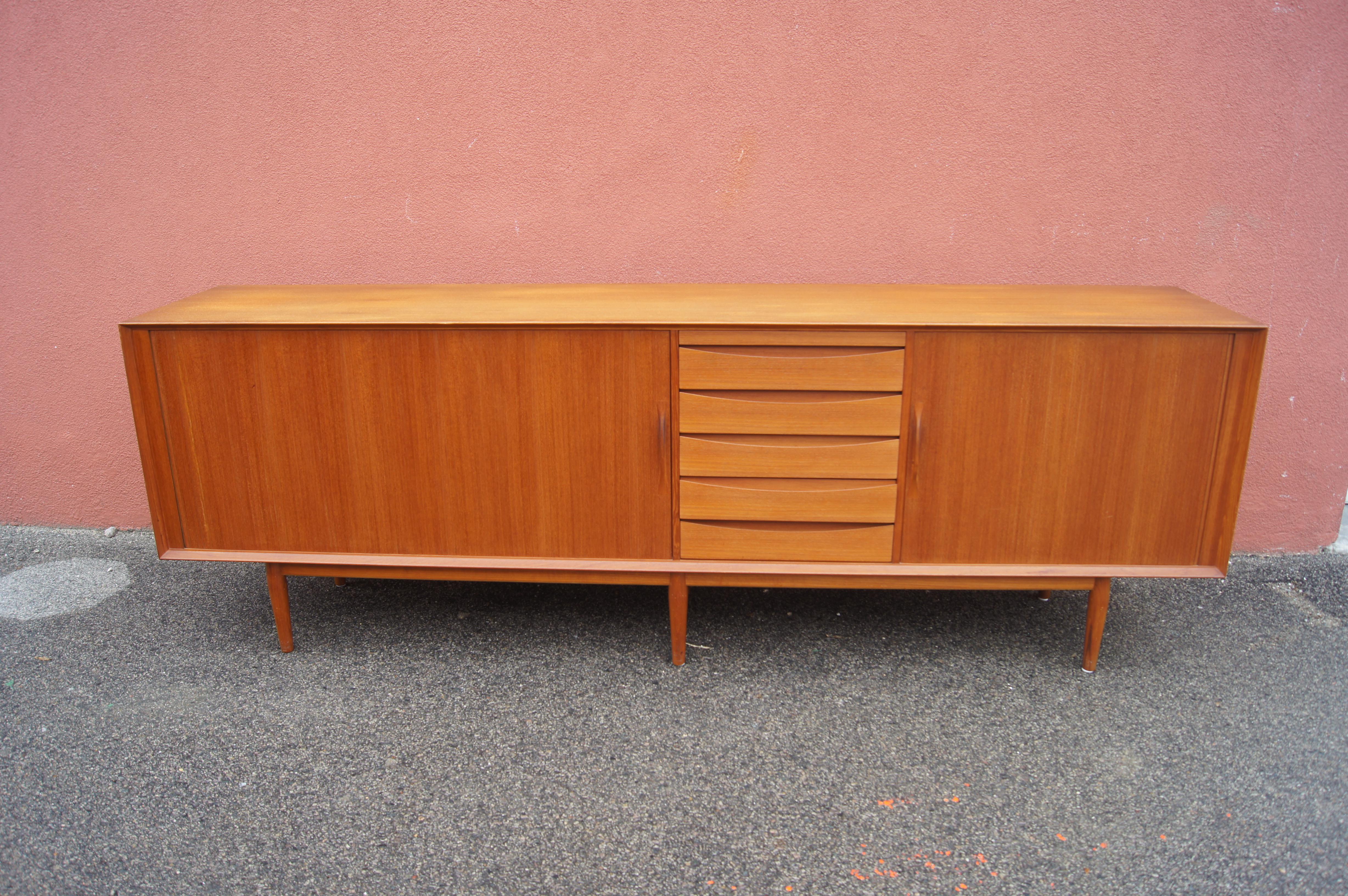 This lovely sideboard, Model 76, by Arne Vodder, places a teak case on a plinth base. In the center, a leather-covered pull-out writing surface sits above five drawers with integrated pulls. On either side, tambour doors with slender vertical pulls