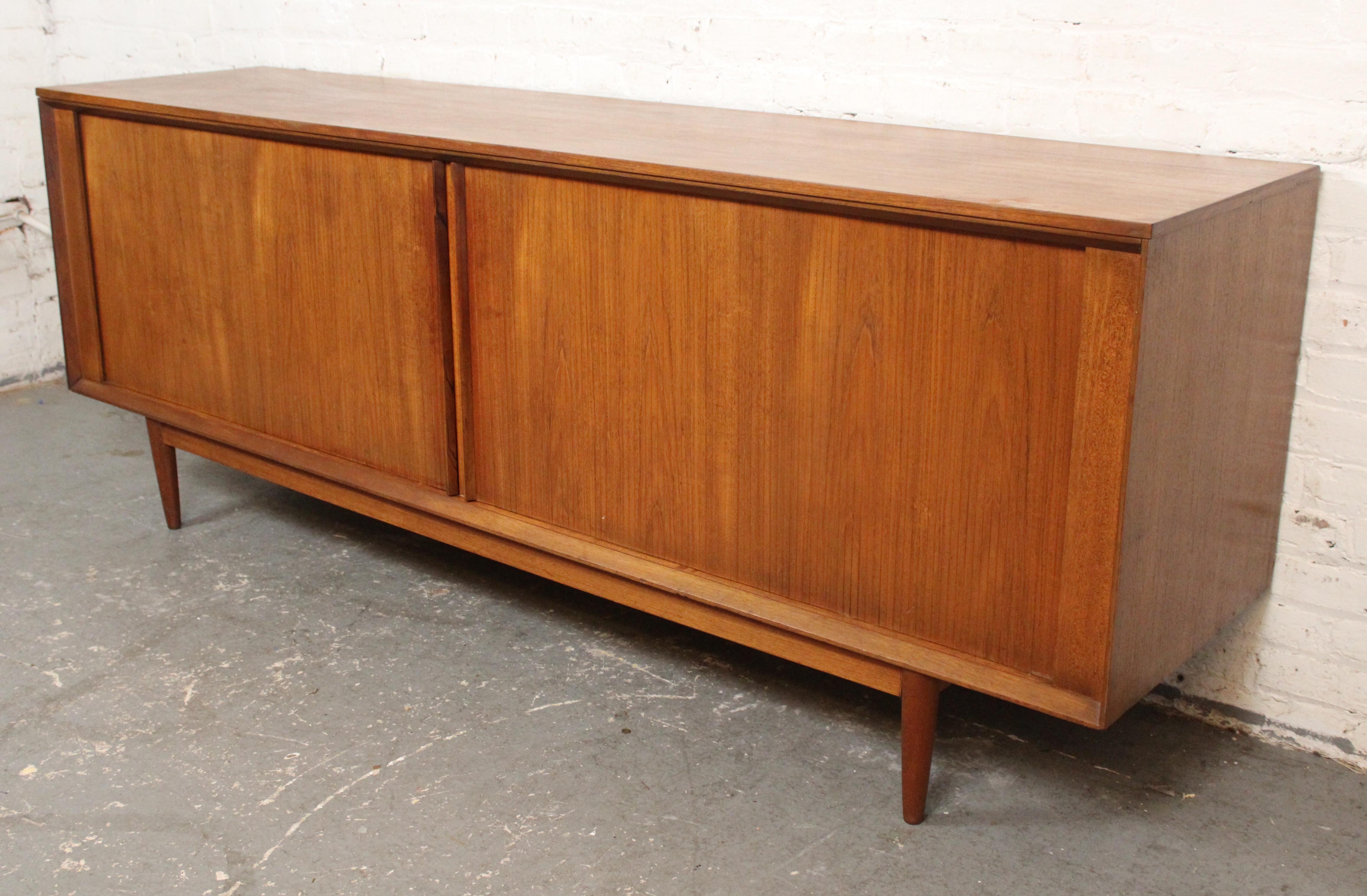 Questa credenza di produzione inglese di Austinsuite Company presenta tutti i segni distintivi della metà del secolo scorso di un server danese. Le venature del legno di teak sono calde, le ante a tamburo si ritirano dietro il mobile, le gambe a