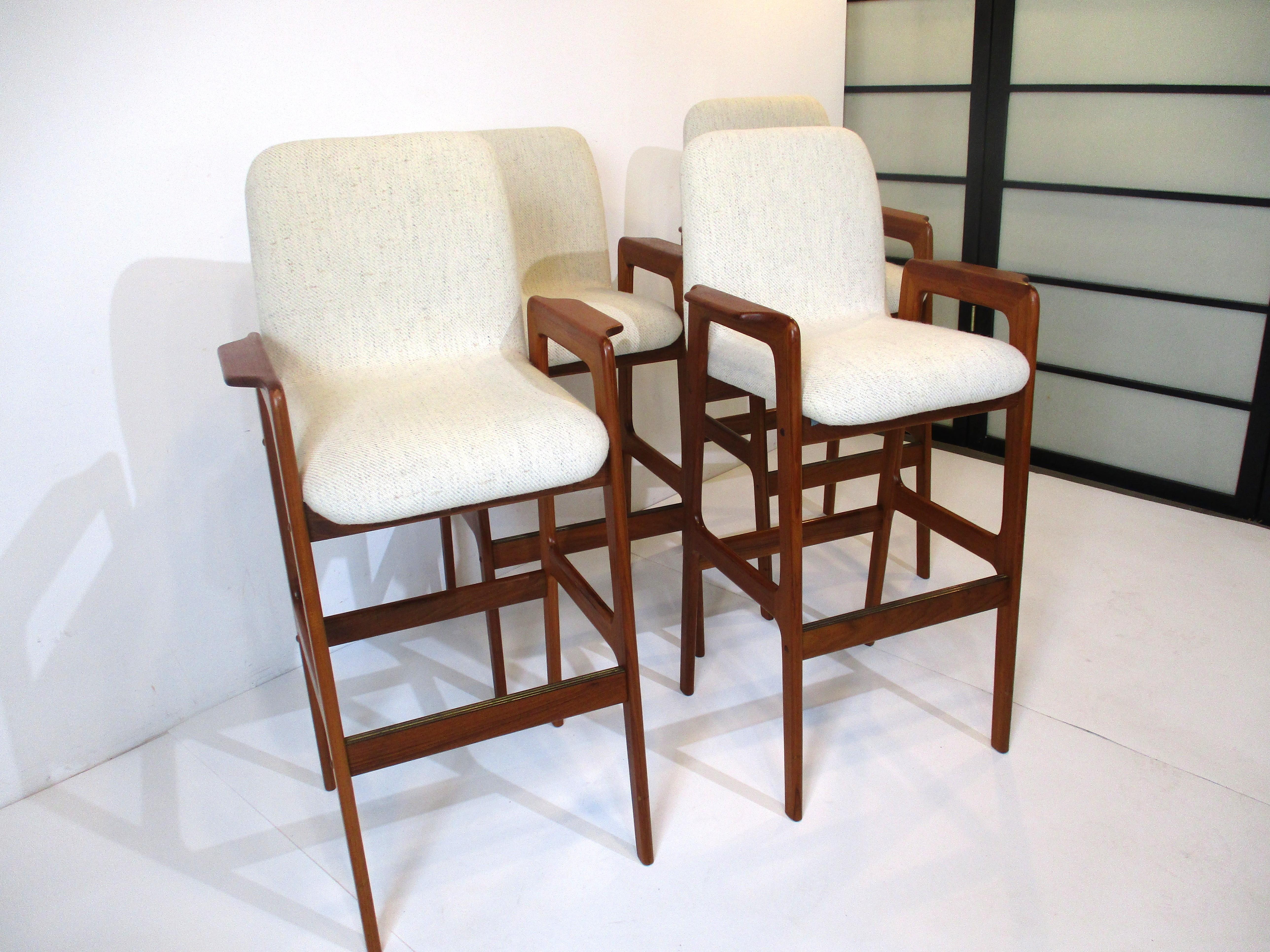 A set of four very comfortable teak wood framed bar stools with nice arm placement. The upholstered seats are in a tight woven darker cream blended fabric with specks of grays and taupes. The foot rest area has a brass trim strip to protect the