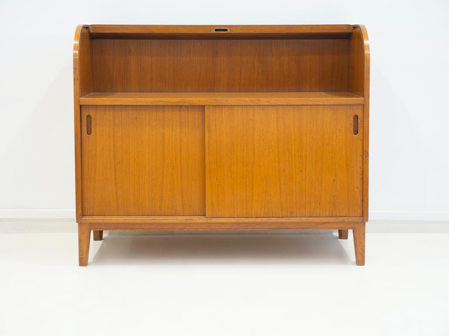 Scandinavian made cabinet veneered with teak. Top part with roll-up flap, bottom with two sliding doors, behind which are storage compartments with a shelf. Key included.

