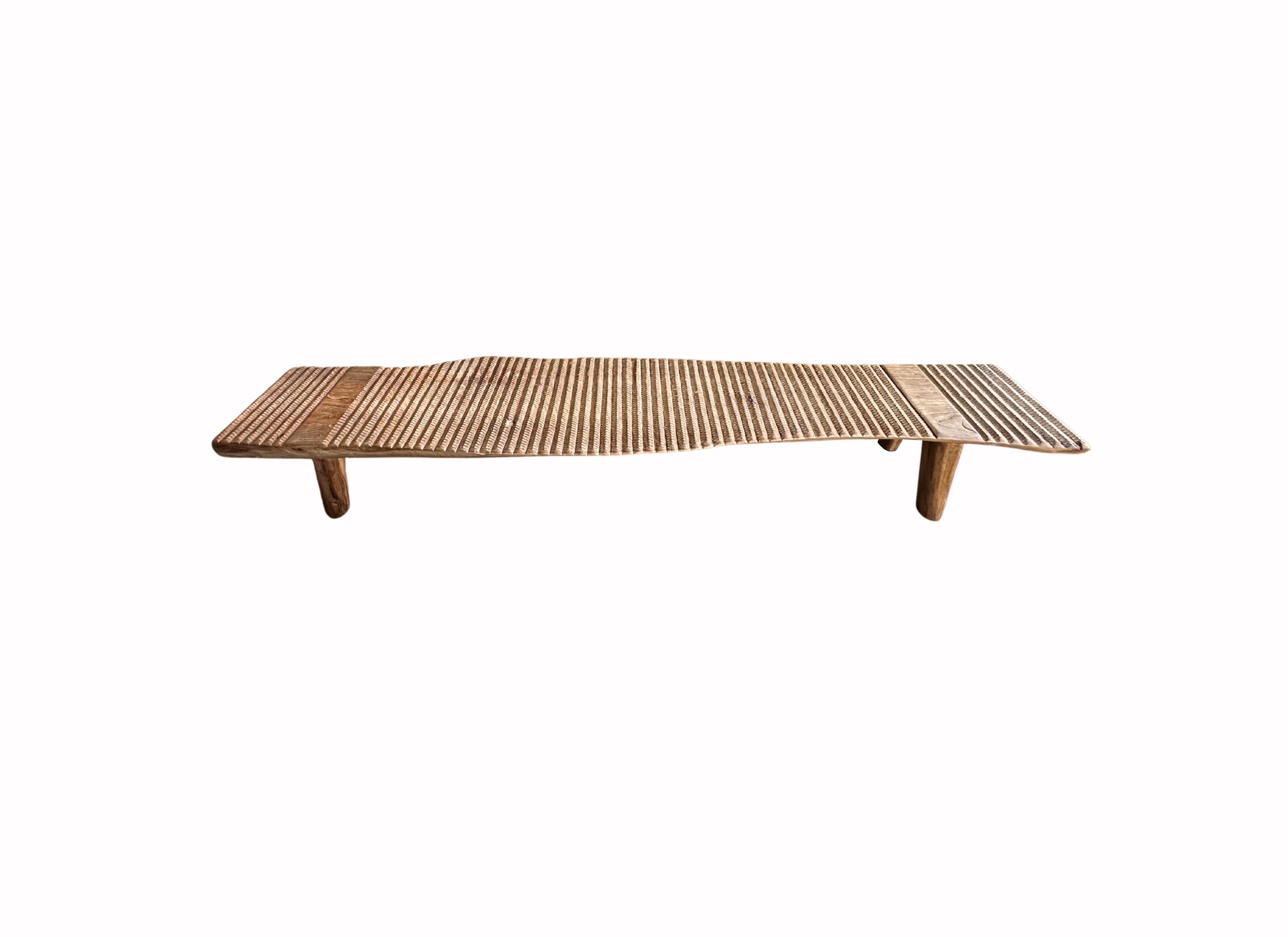 A wonderfully sculptural teak wood bench with carved detailing on its seat. This teak wood bench features a wonderful organic form with a mix of wood textures and shades. Elevated by four slender teak wood legs, the seat was carved from a single