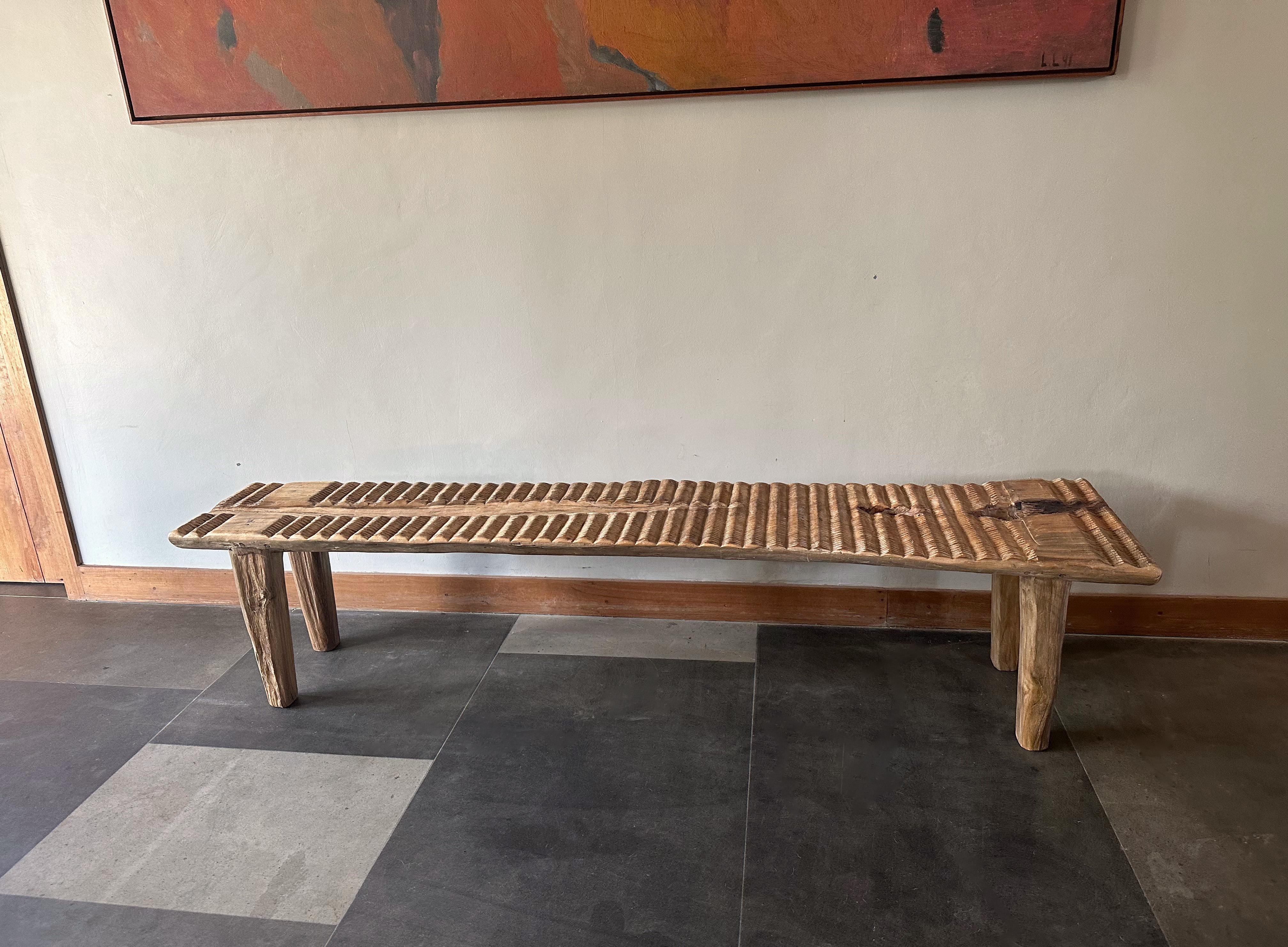 A wonderfully sculptural teak wood bench with carved detailing on its seat. This teak wood bench features a wonderful organic form with a mix of wood textures and shades. Elevated by four slender teak wood legs, the seat was carved from a single