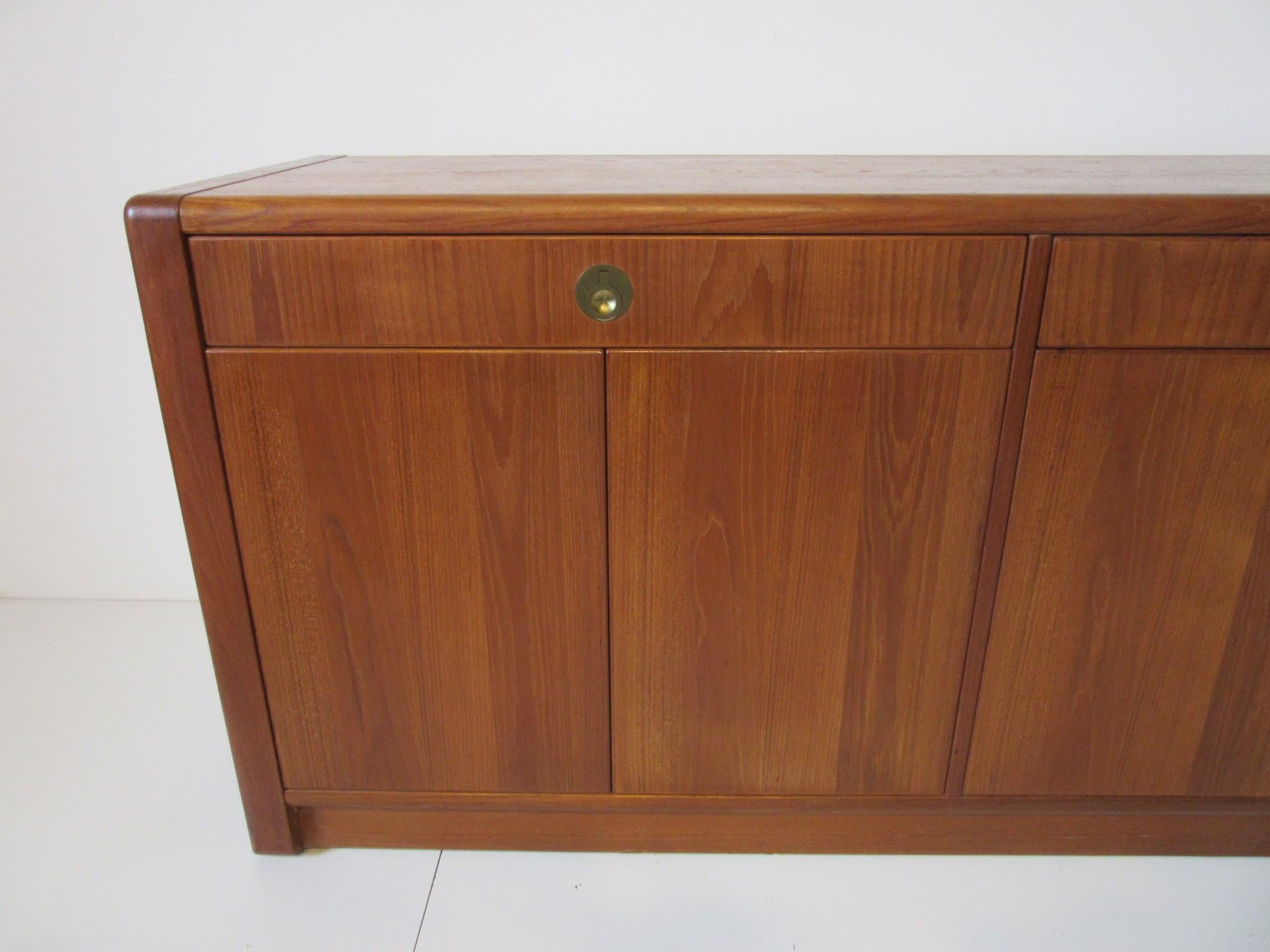 A medium dark toned teak server / chest / credenza with inset finger joint construction, four doors that have push touch latches and adjustable shelves. The two upper drawers have heavy brass pulls manufactured by D - Scan Singapore from the Captain