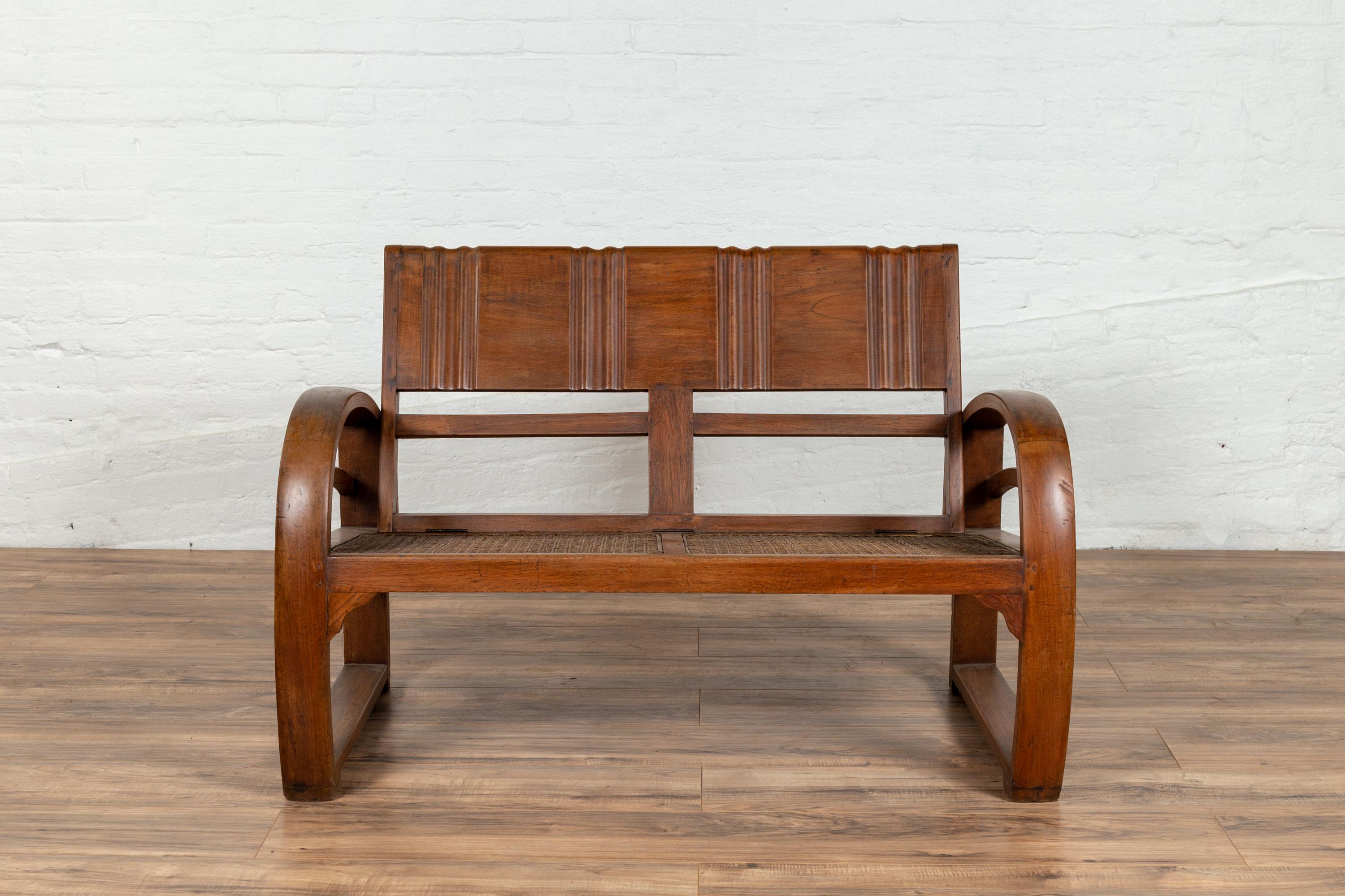 An antique Indonesian teak wood settee from the early 20th century, with cane two-seater and looping arms. Found in Madura, this early 20th century teak settee features a rectangular back with reeded accents that conveniently folds to rest on the