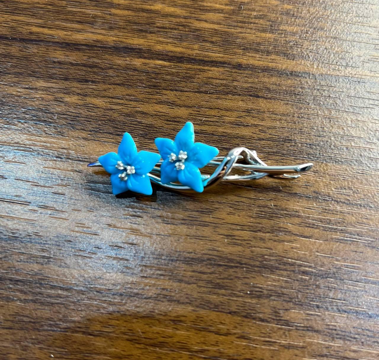 This charming brooch features two vibrant blue turquoise flowers, each with a delicate cluster of shimmering accent diamonds at their centers. The flowers are set against a sleek, intertwined 14k White Gold base, creating a harmonious blend of color