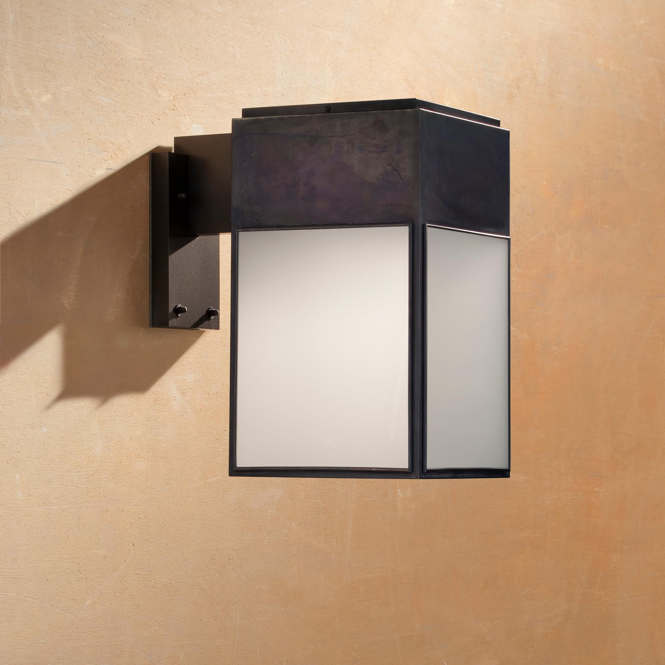 Eclectic wall light in brass with outside fitted clear or frosted glass, closed top and open bottom. The closed upper part is also the base for the electrical equipment. Wall bracket in black lacquered aluminium with forcible transit. For outdoor