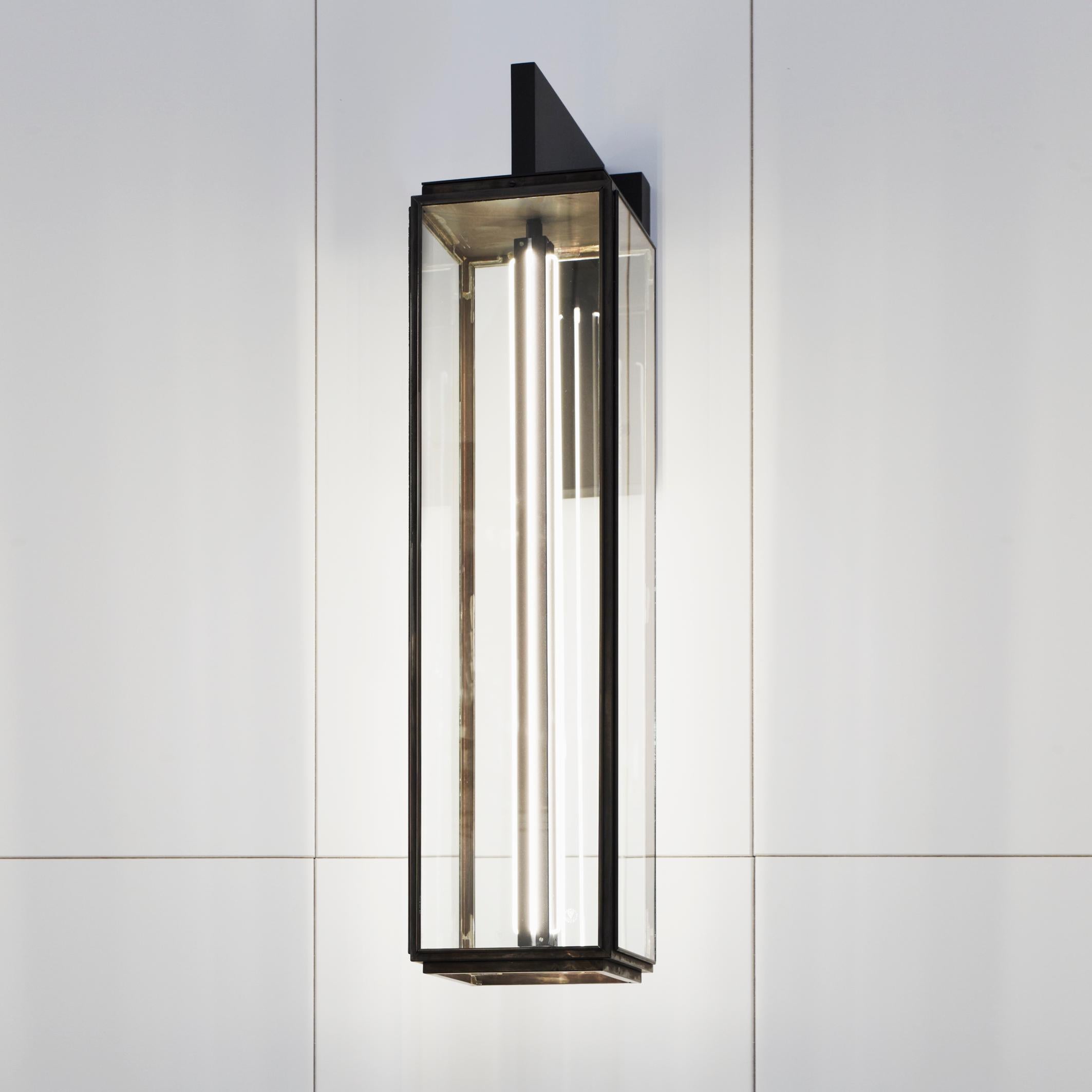 Eclectic wall light in dark bronze with outside fitted clear glass, closed top and open bottom. Wall bracket in bronze lacquered inox with forcible arm. Central lamp holder with four cold cathode lamps. For indoor use only (IP20).

Lamp cold