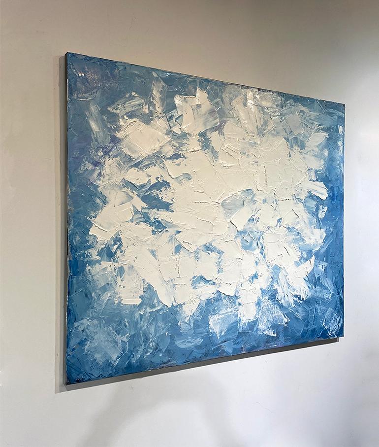 This large abstract painting by Teodora Guererra features shades of blue,  pops of lavender and white for its palette. Paint has been applied with various palette knives layering like frosting with a thick textured surface.  The painting has a