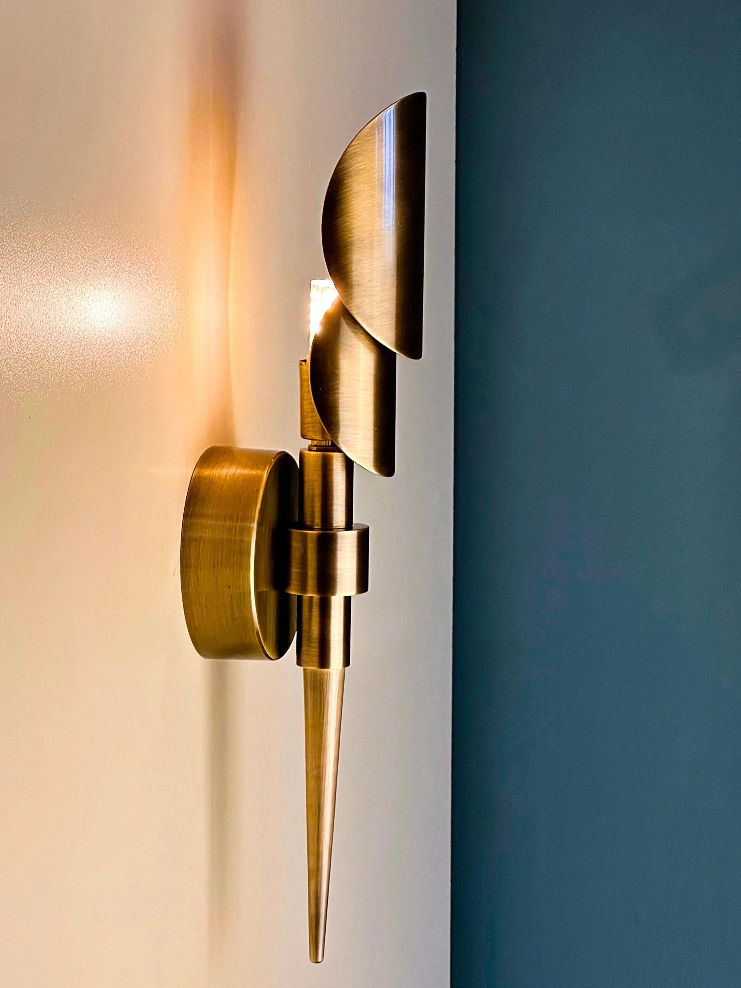 El aplique de pared de latón TEREQUE es una sorprendente encarnación de la elegancia moderna de mediados de siglo, elaborado totalmente a mano para celebrar tanto la forma como la función. Su núcleo es un elegante tubo cónico de latón que sujeta con