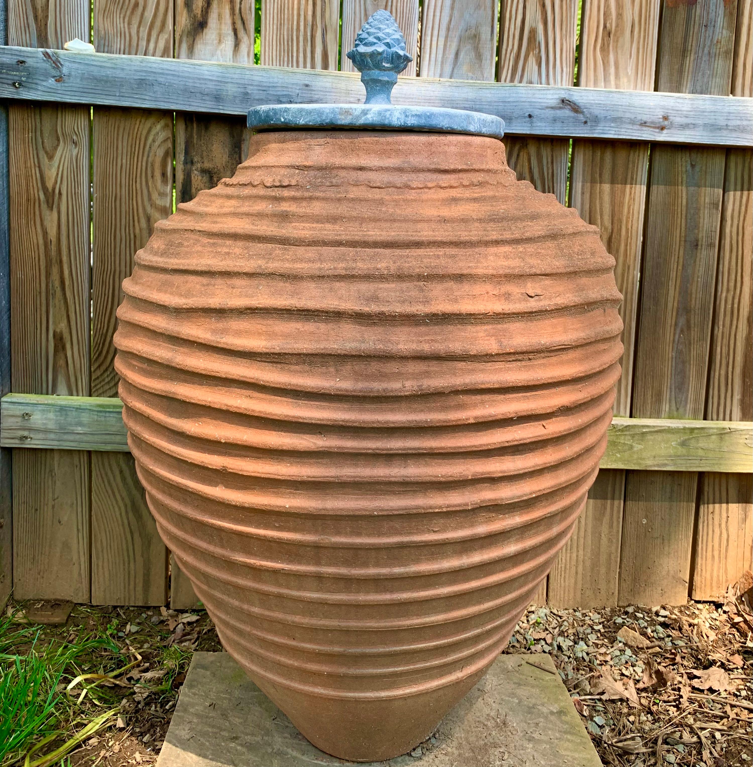 A very large spiral fluted terracotta pot garden ornament, with a tapered and slightly flared neck. The pot is topped by a distinctive ornamental lead cover surmounted by a cast lead acanthus and pinecone finial. 20th century. Flat on the bottom.