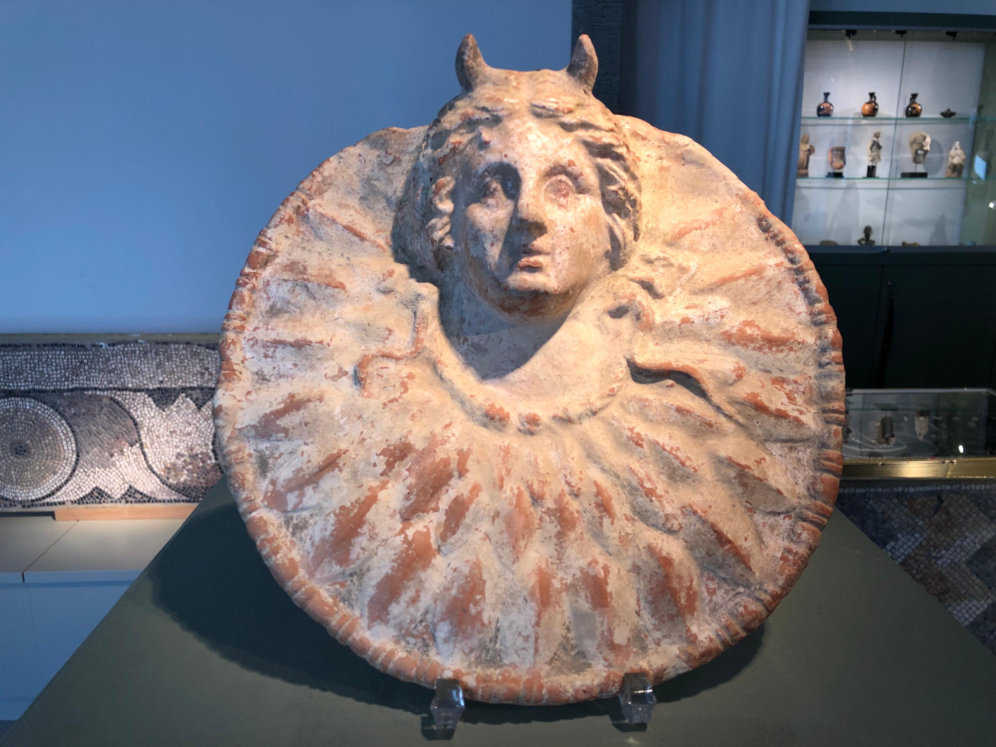 Round disc framed by an astragal band with almost plastic winged head of Medusa. Around her neck Medusa wears a torque and snake tails. The head is surrounded by scales indicating that the tondo represents the aegis respectively the shield of