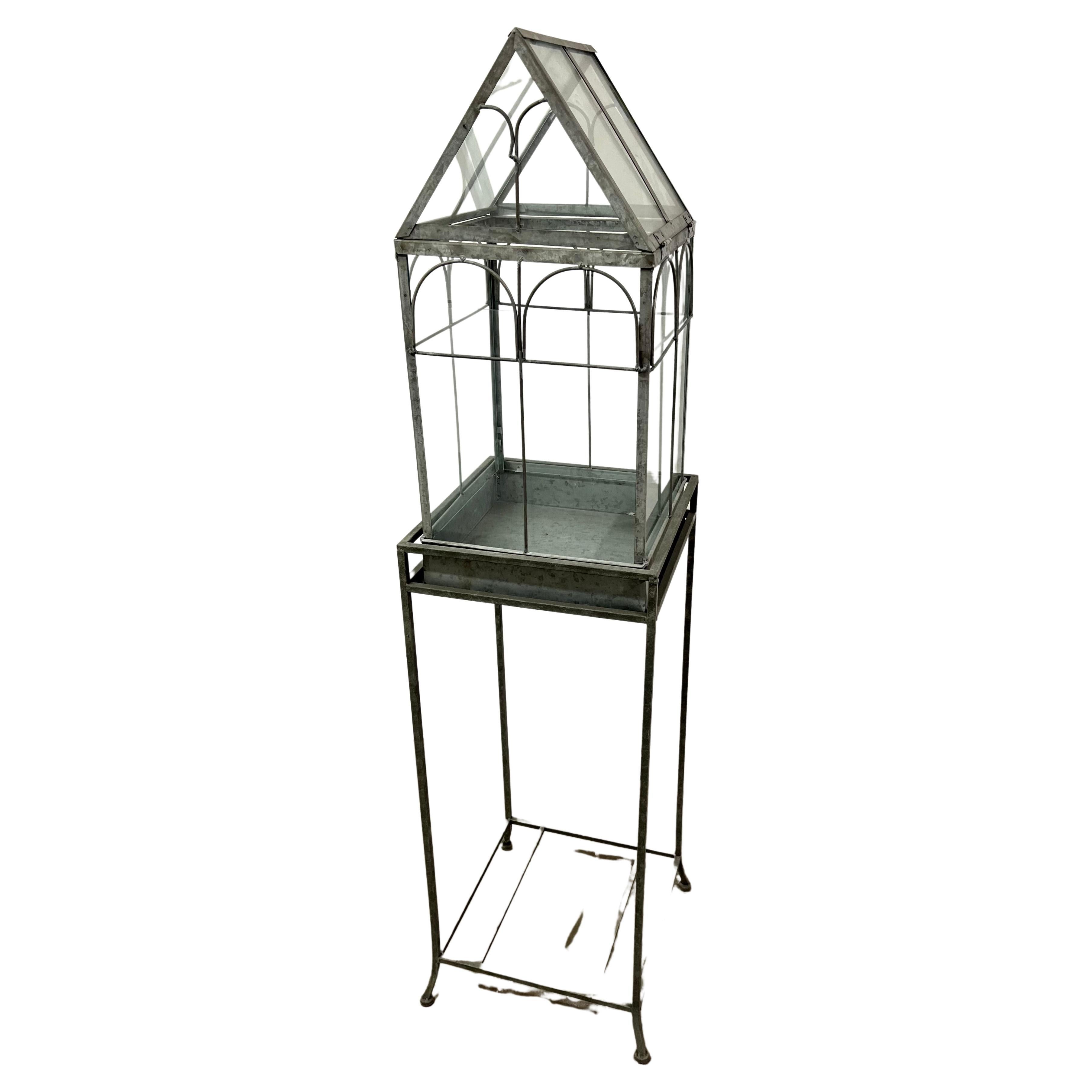 A glass house sits atop an open metal tray pedestal with ball feet. This piece opens from the top. 

This piece is functional indoors or outdoors for creating humid environments for plants. The original metal has been gorgeously patinated over