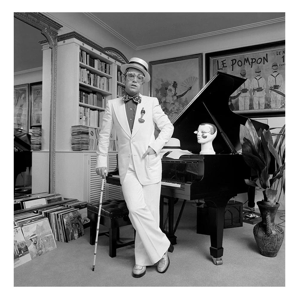 Elton John est photographié dans sa tenue excentrique reconnaissable à côté du piano dans sa maison de Windsor, en Angleterre, en 1974. Cette image a été prise dans le cadre de la réalisation de la pochette du onzième album du musicien, Elton John's