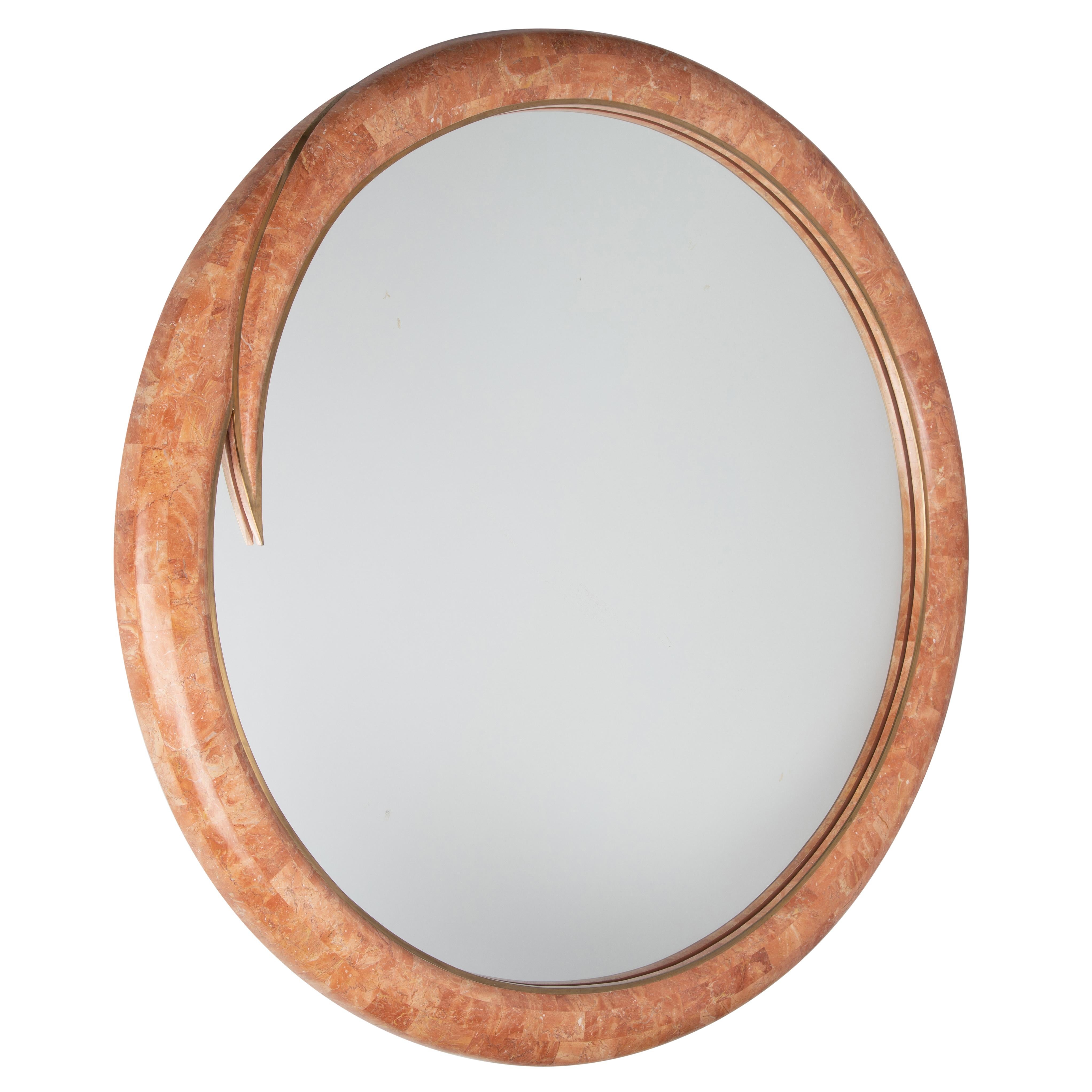 This Maitland Smith mirror features a rounded wooden frame covered in coral-color tessellated marble with an inner band of brass trim. The circular form resolves in a whimsical 