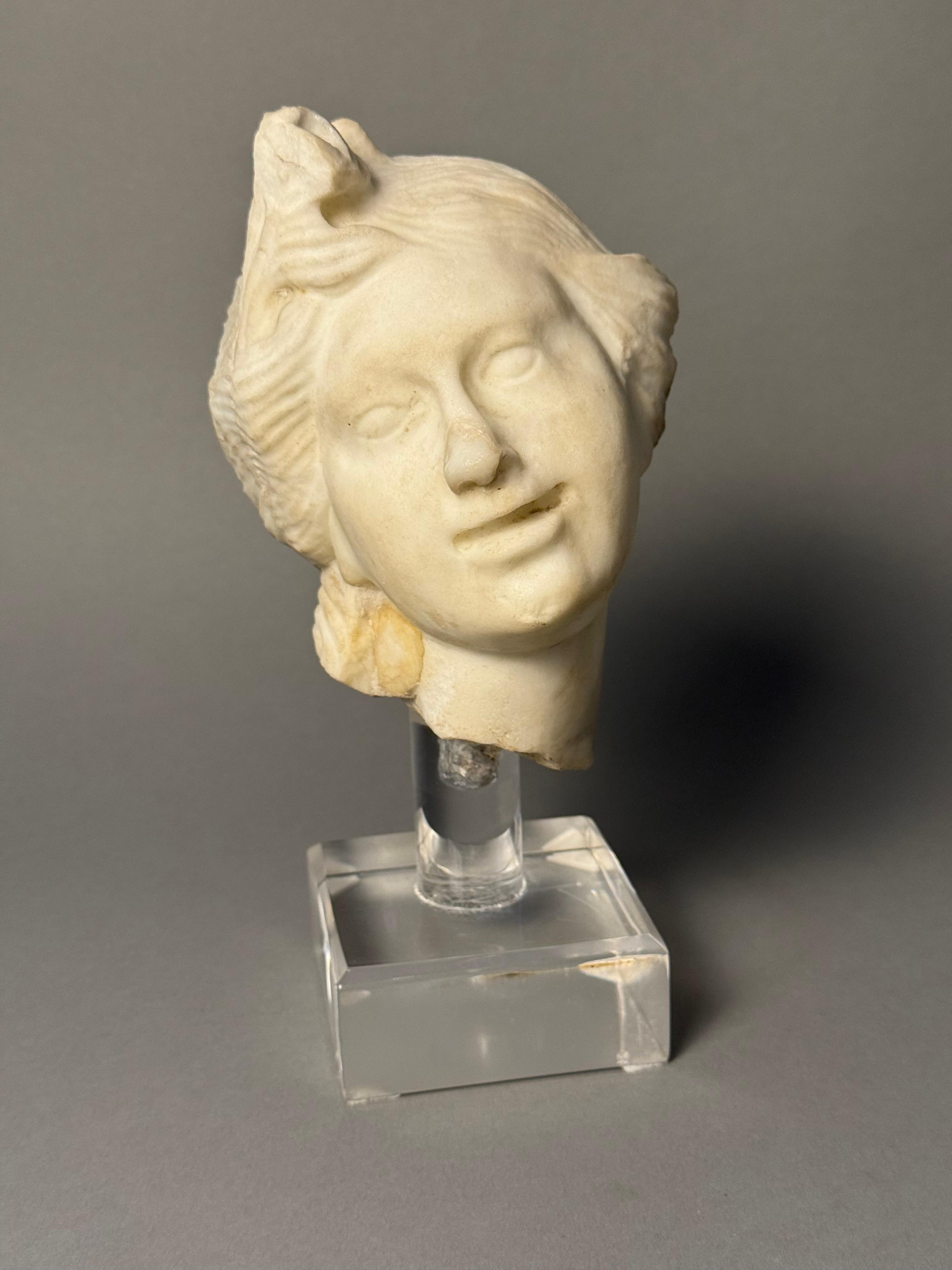 Testina in marmo neoclassica del XVIII secolo che raffigura la testa e parte del collo di una figura femminile. L'acconciatura così elaborata richiama l'arte greca o romana. La figura ha un'espressione serena e sembra abbozzare un leggero