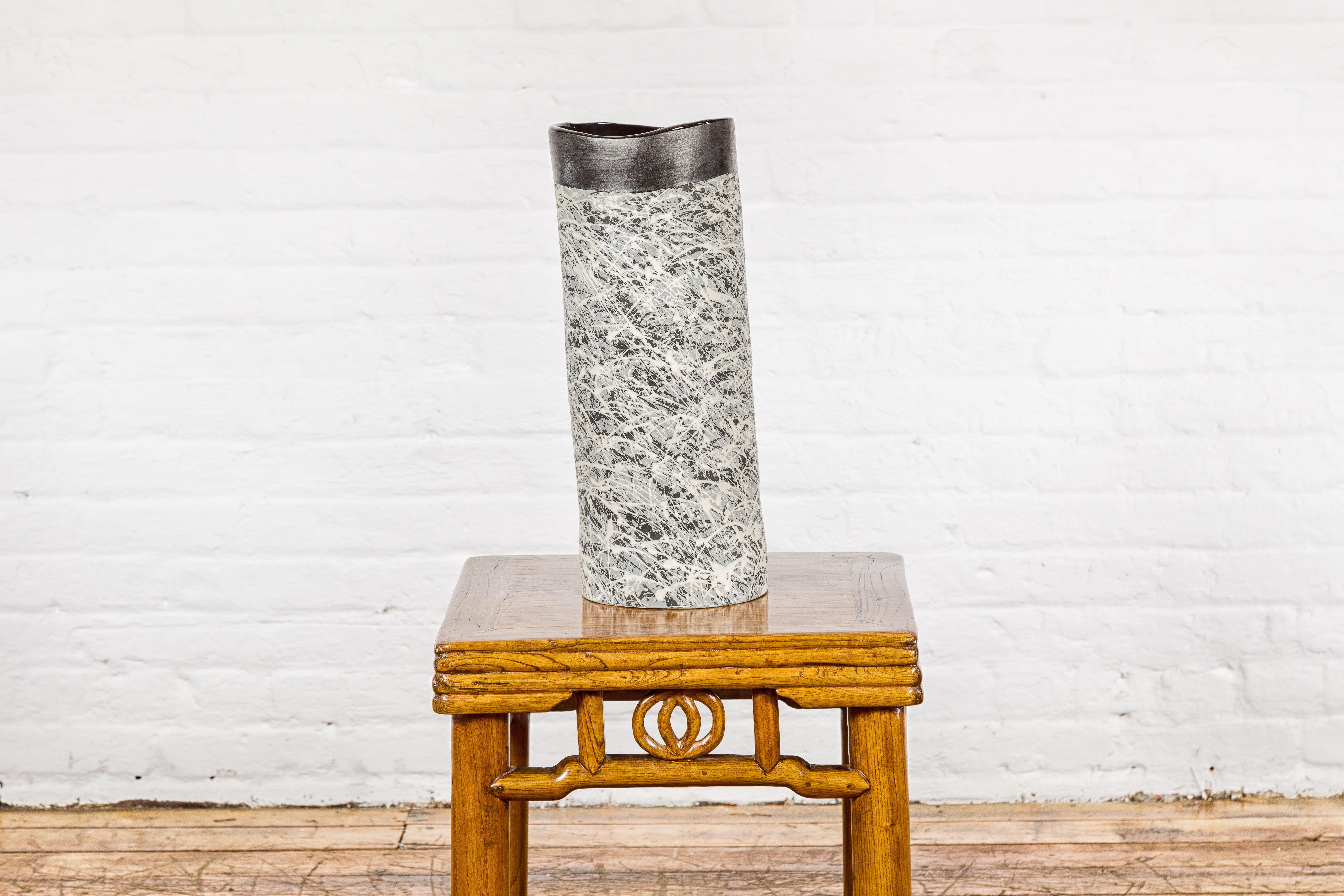A textured black, gray and white spattered ceramic vase. This ceramic vase captivates with its oblong silhouette and an arresting spatter of black, gray, and white, creating a dynamic abstract pattern. The textured surface adds depth and intrigue,