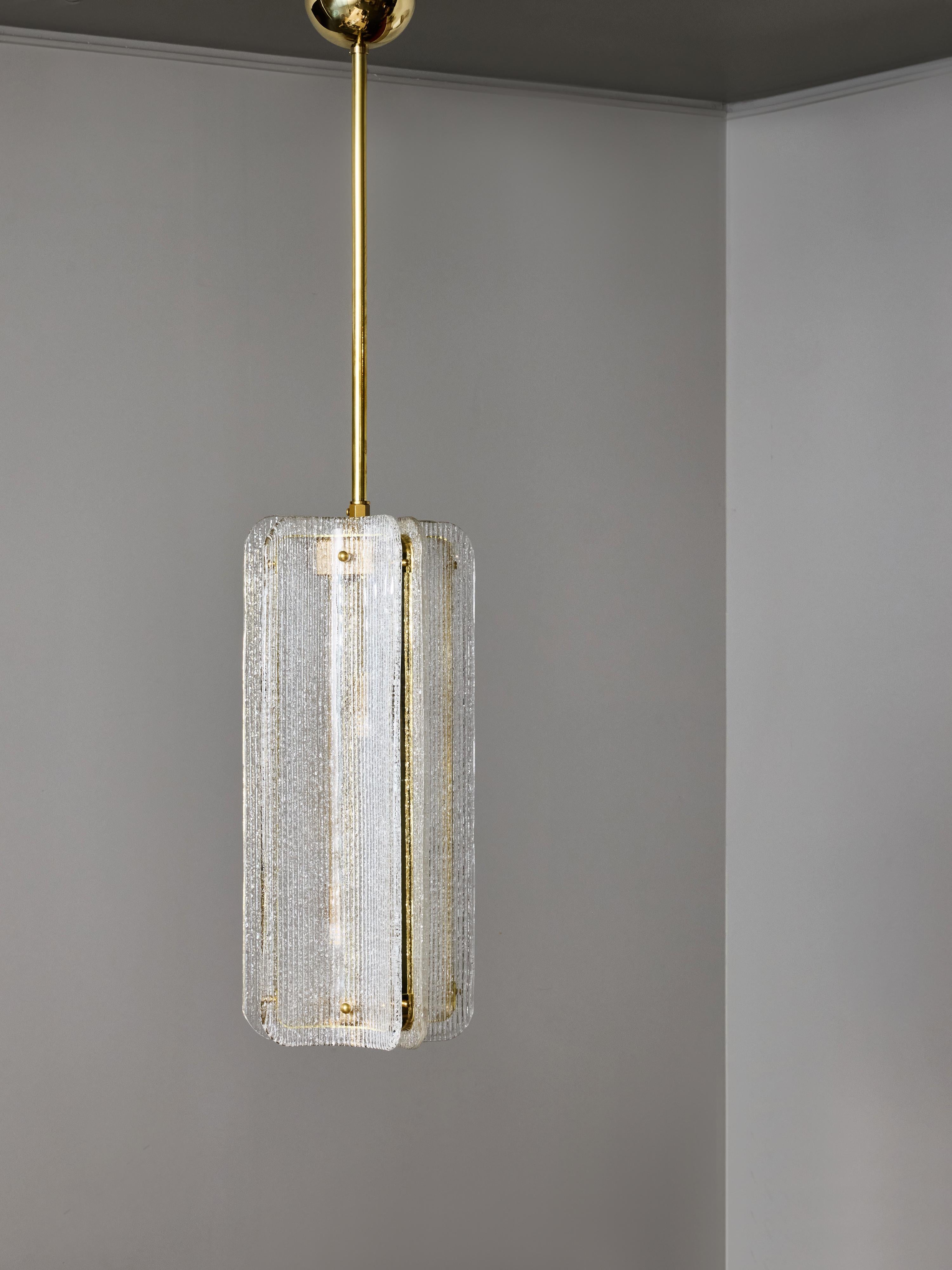 Una sofisticada lámpara colgante moderna, con un conjunto escultural de paneles de cristal texturizado montados alrededor de una estructura central de latón. La lámpara se compone de cuatro elementos de cristal curvados verticalmente, cada uno con